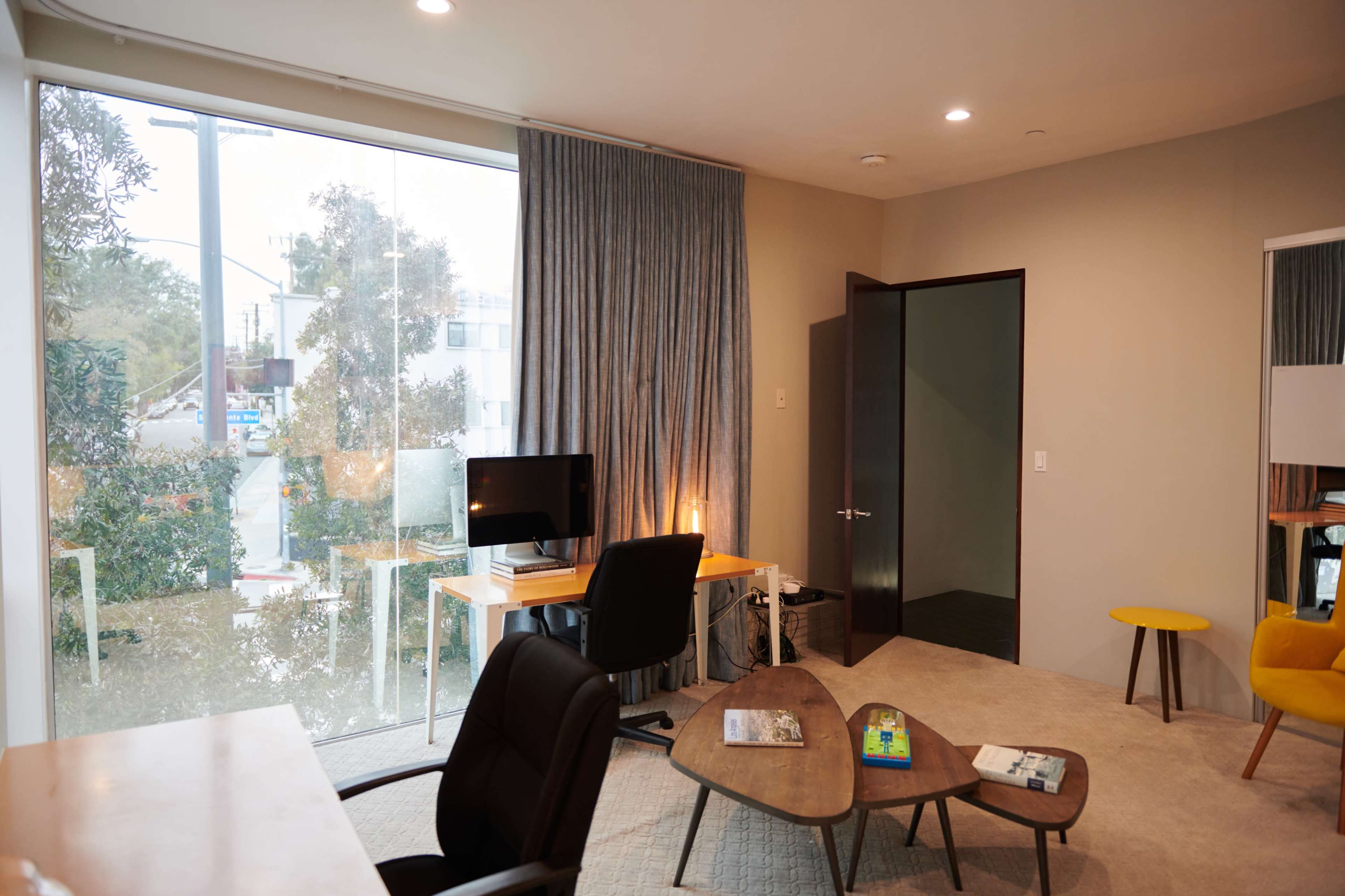 A modern workspace features a desk with a computer, a chair, and a window with curtains, alongside a small coffee table and a yellow accent stool.