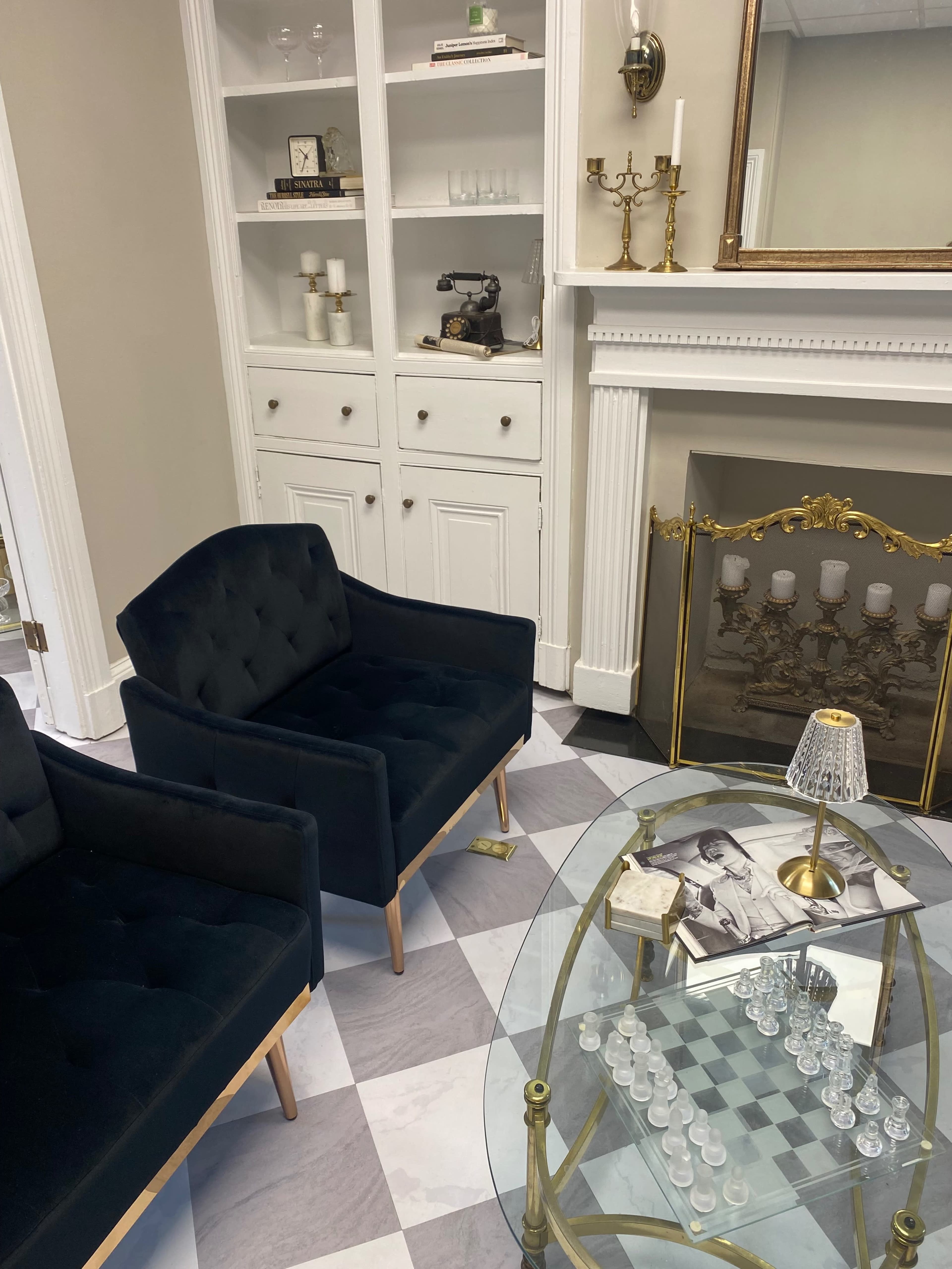 The image shows a cozy room featuring two dark blue chairs, a glass coffee table with a chess set, a vintage sewing machine on a shelf, and elegant decor elements around a fireplace.