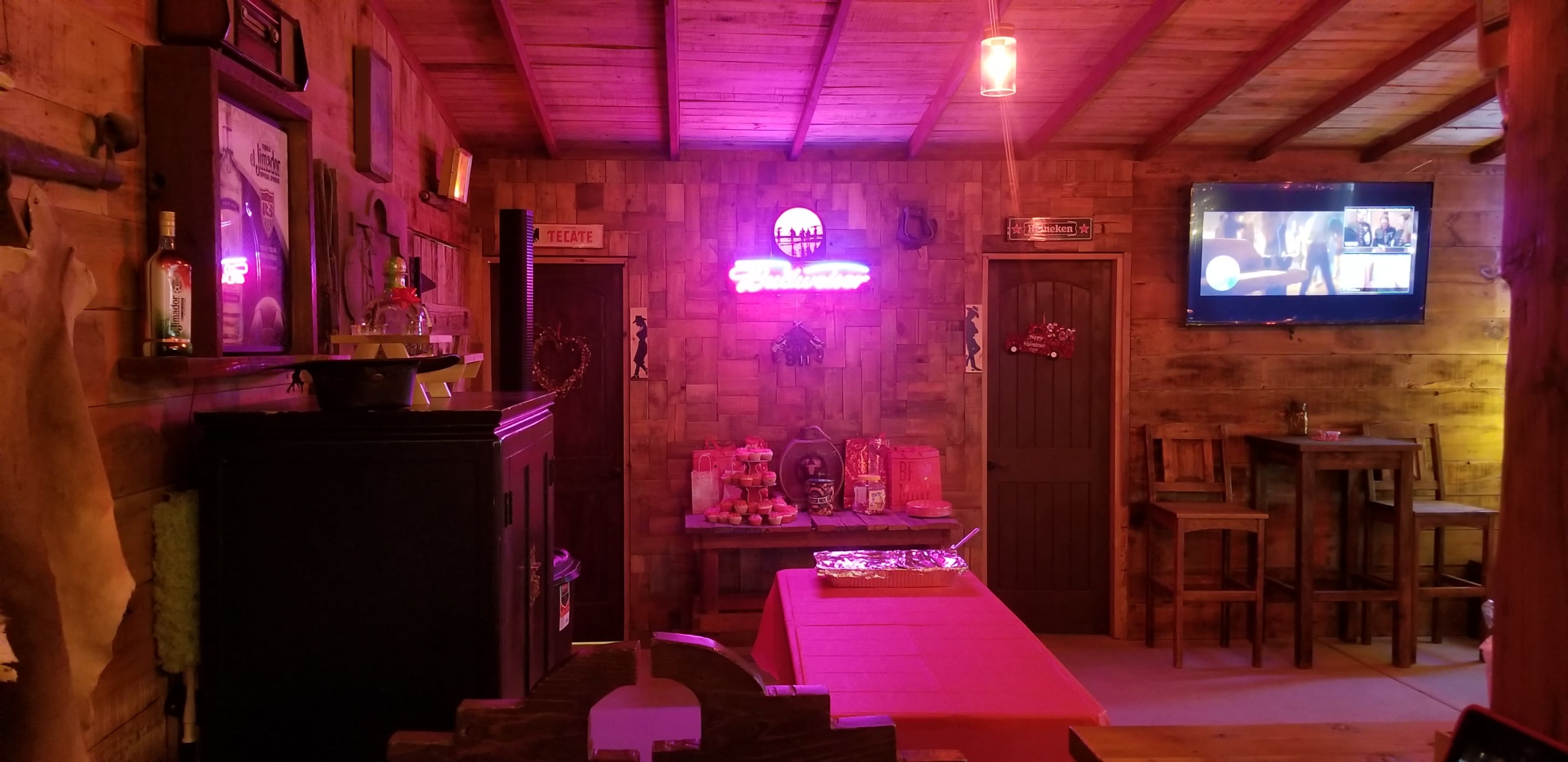 The image shows the interior of a rustic bar with wooden walls, a neon sign, a television, and tables arranged for seating.