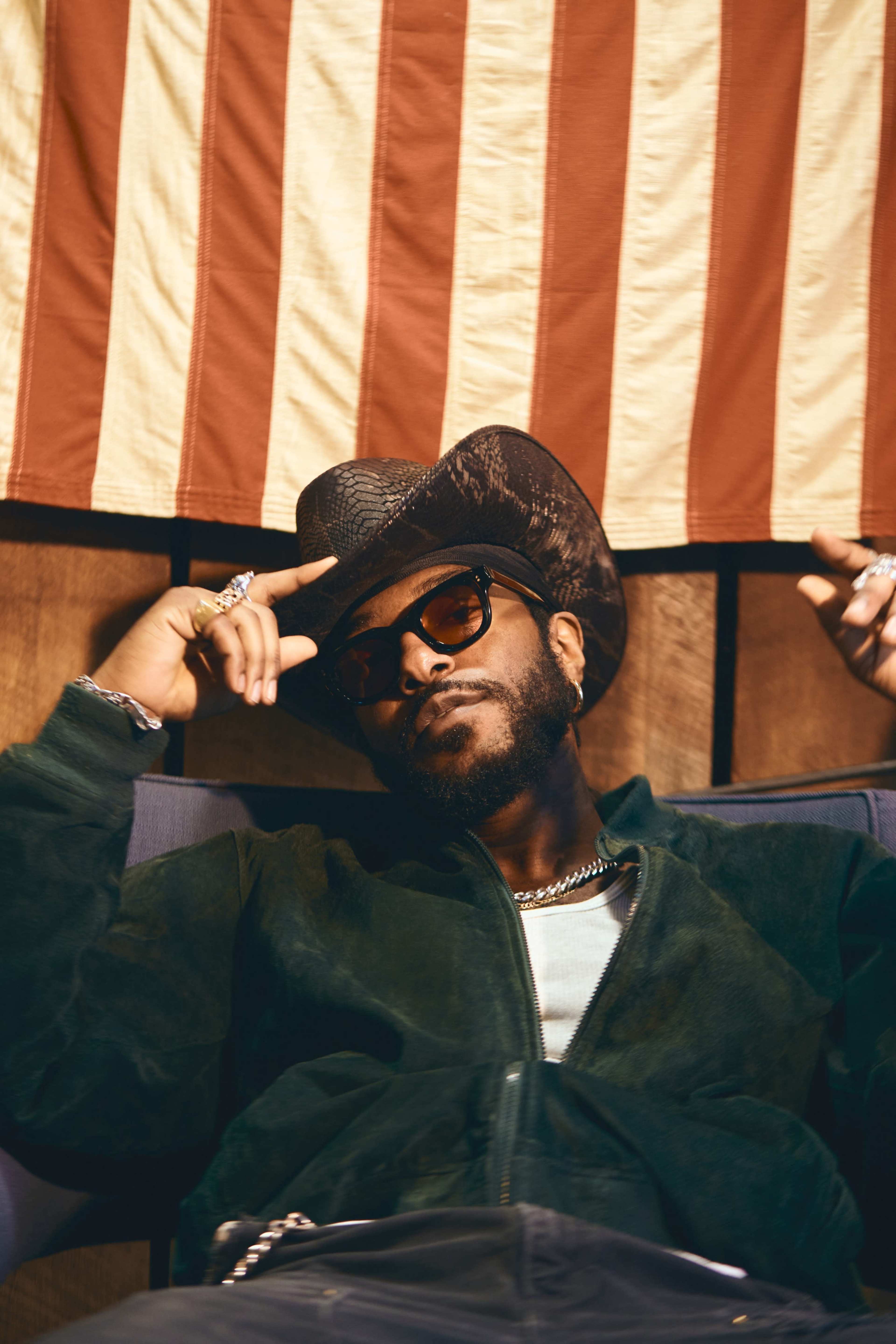 A person wearing sunglasses and a cowboy hat is reclining against a wall adorned with an American flag.
