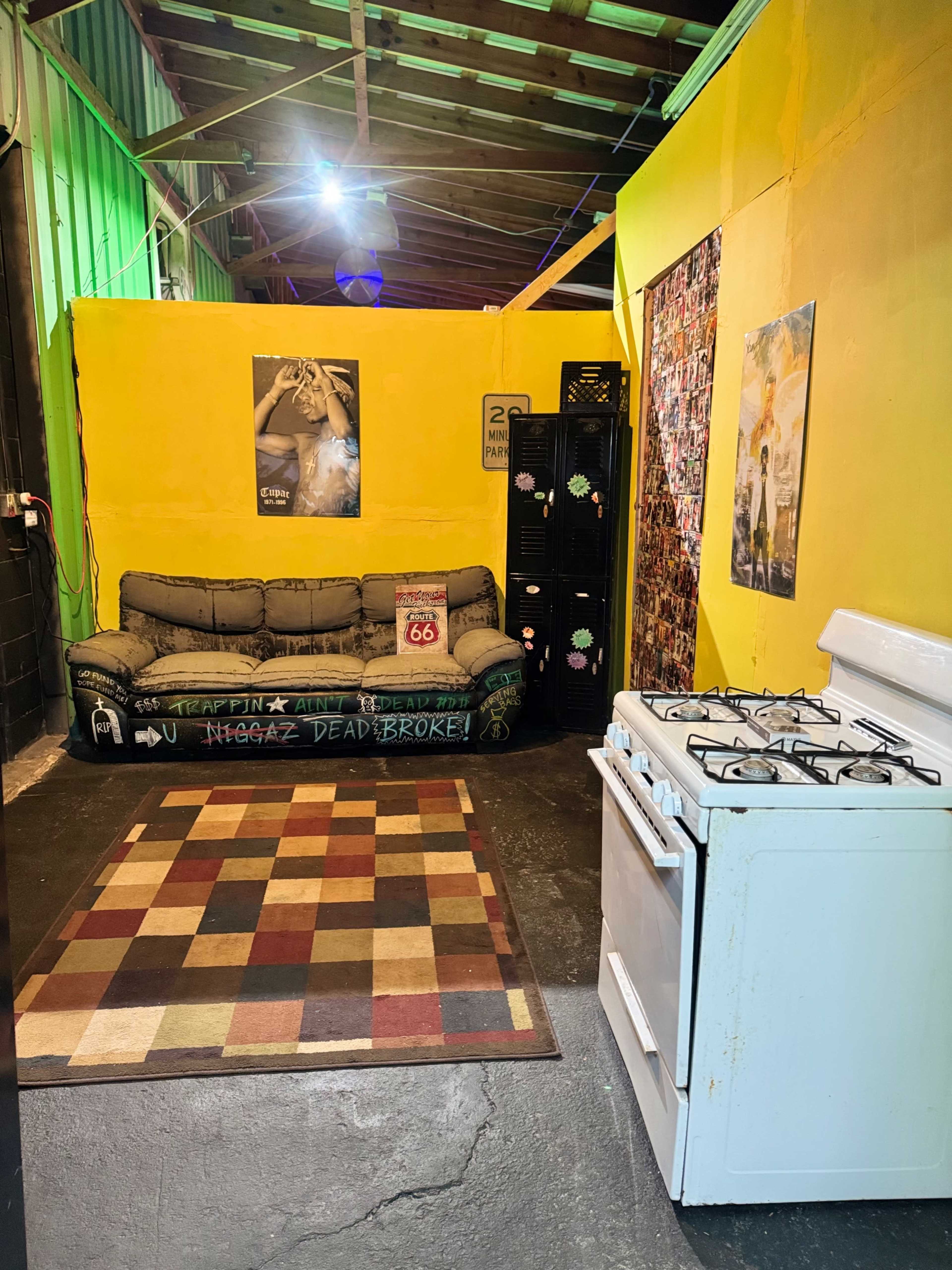 The image shows a small, yellow-walled room with a couch, a gas stove, and a patterned rug on the floor.