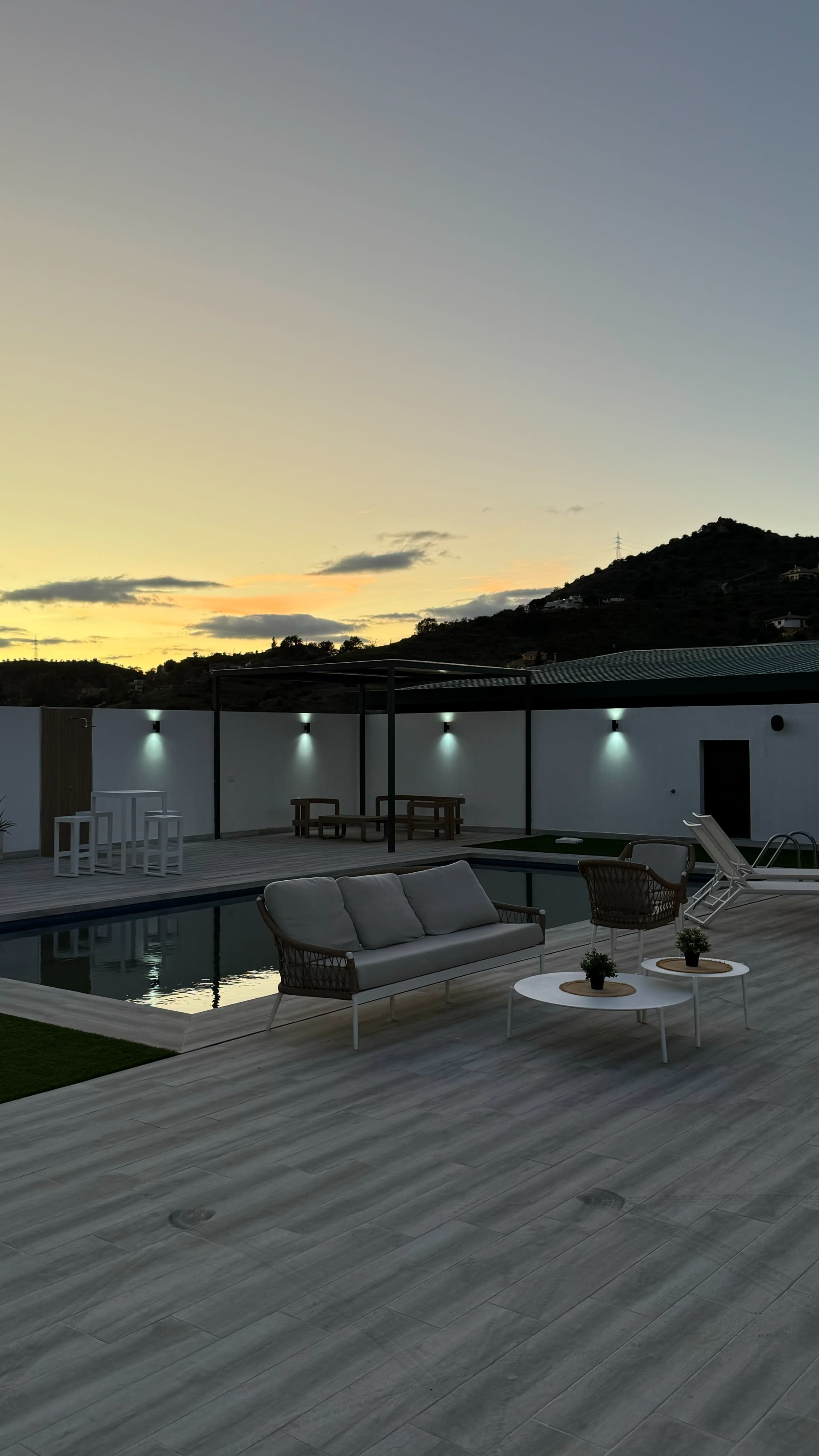 The image shows a modern outdoor space with a swimming pool, lounge furniture, and a sunset backdrop over a mountainous landscape.