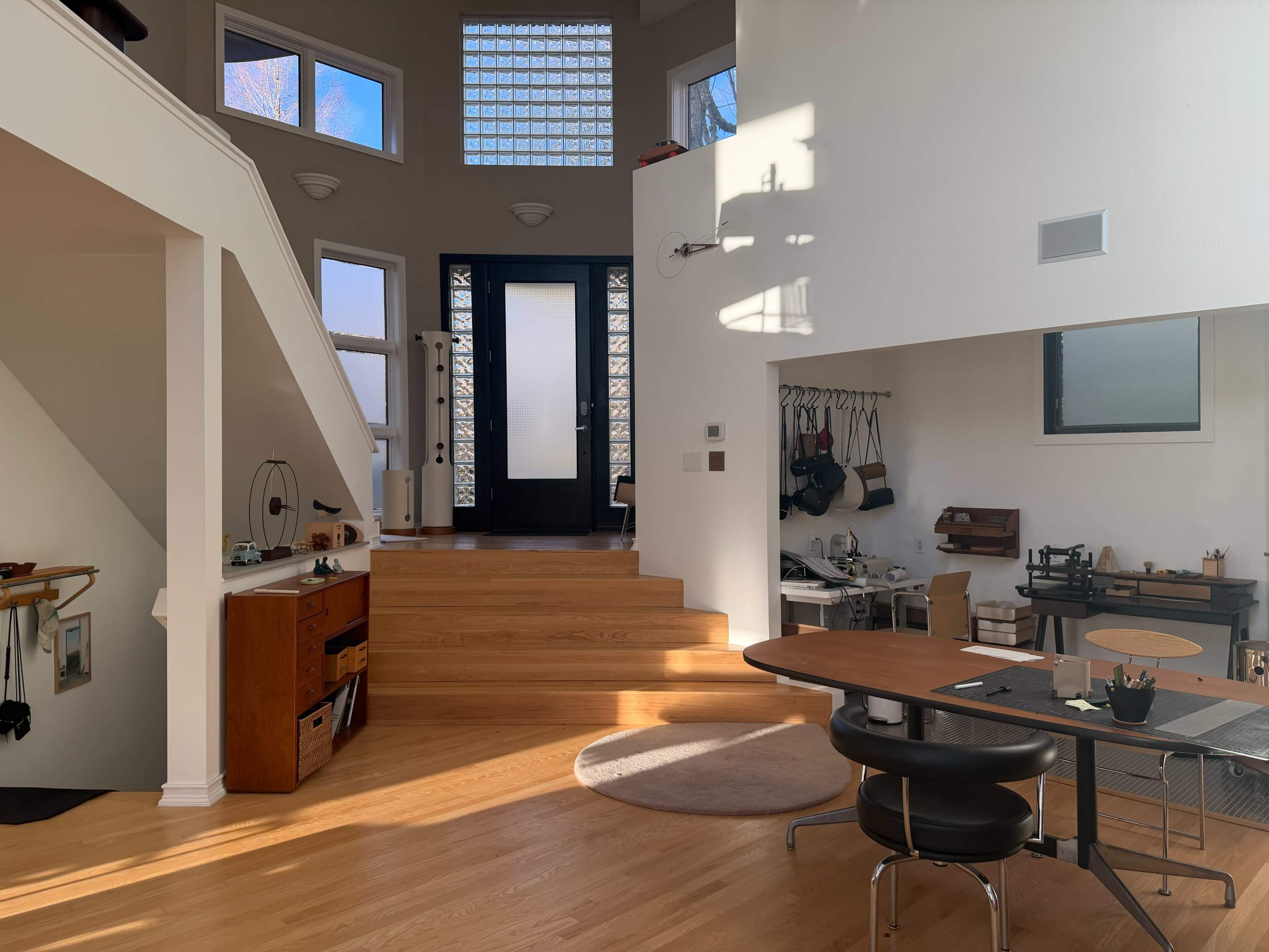 The image shows a modern, bright interior space with a staircase leading to a black door, a large window, and a dining table with varying furniture and decor.