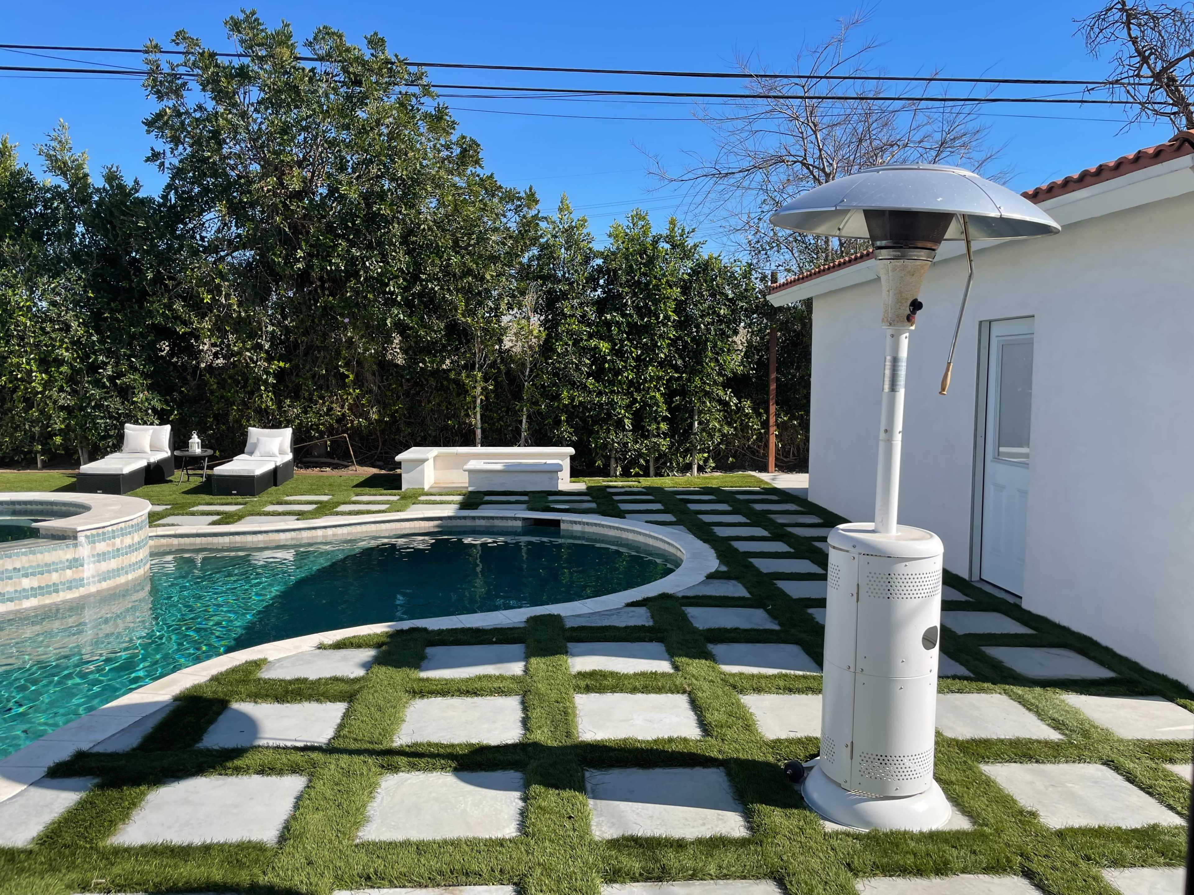 A backyard scene featuring a swimming pool surrounded by green grass and stone pavers, with patio heaters and lounge chairs beside it.