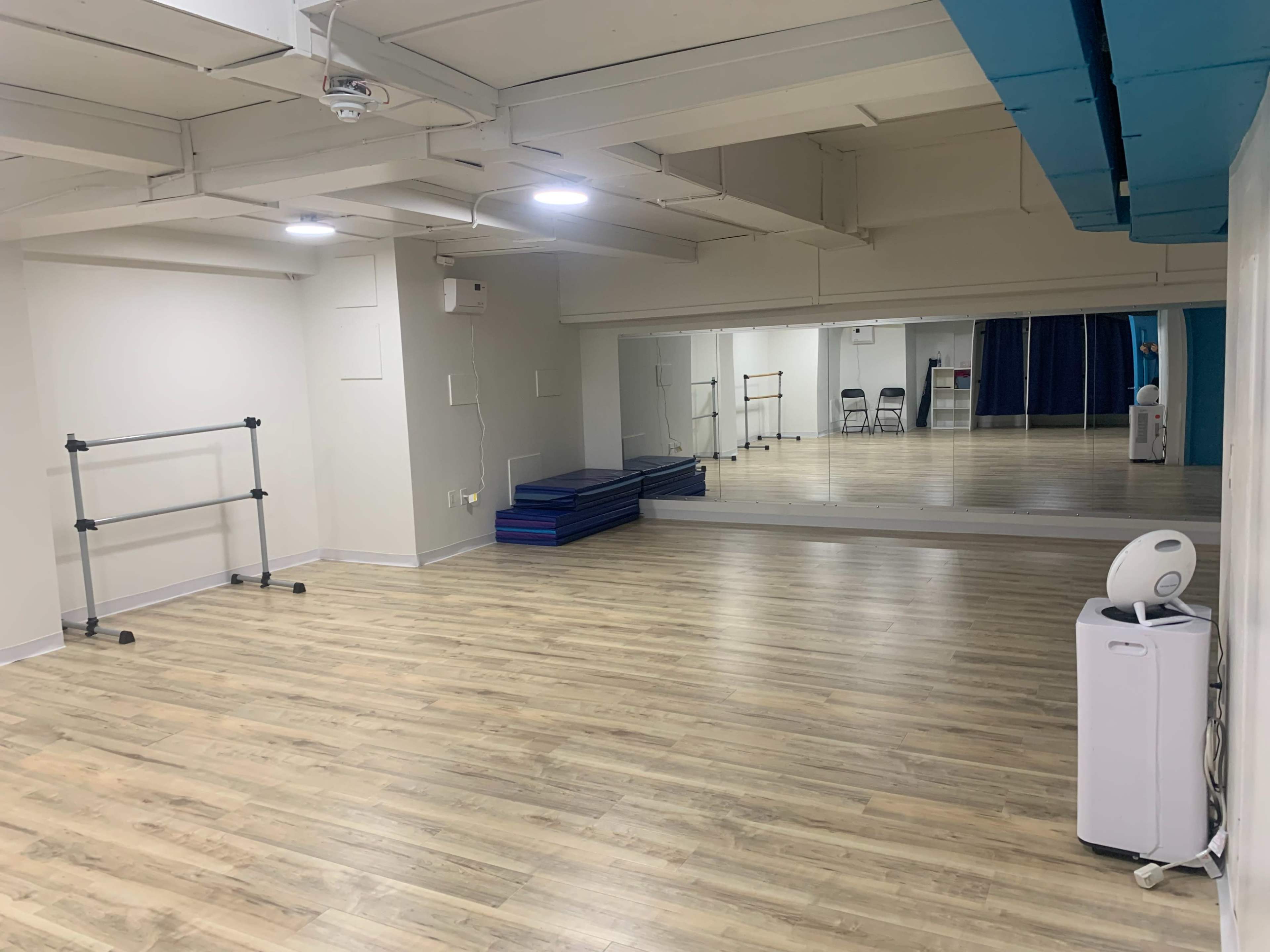 The image shows a spacious, well-lit studio with wooden flooring, a large mirror on one wall, and some exercise mats neatly stacked in one corner.