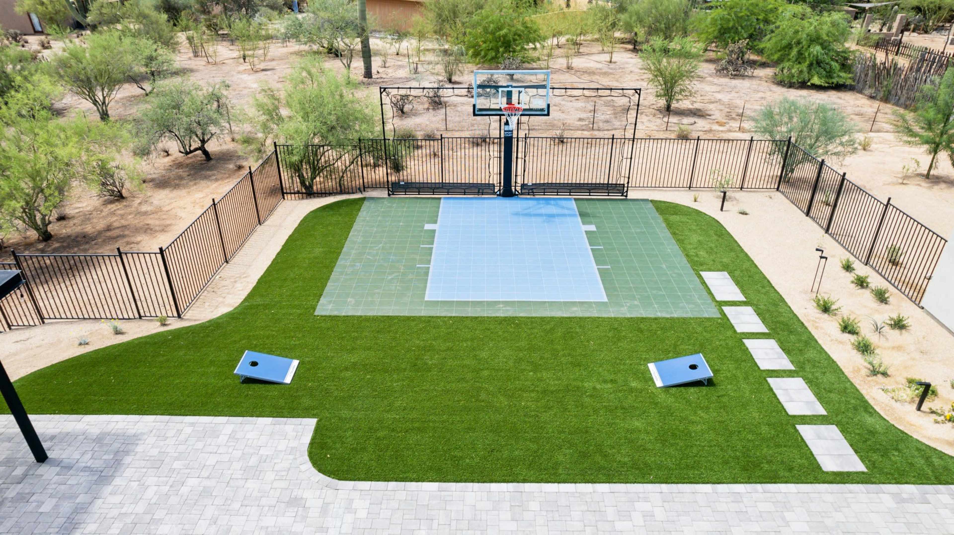 The image shows an outdoor basketball court with a hoop, surrounded by green turf and gravel pathways in a desert landscape.