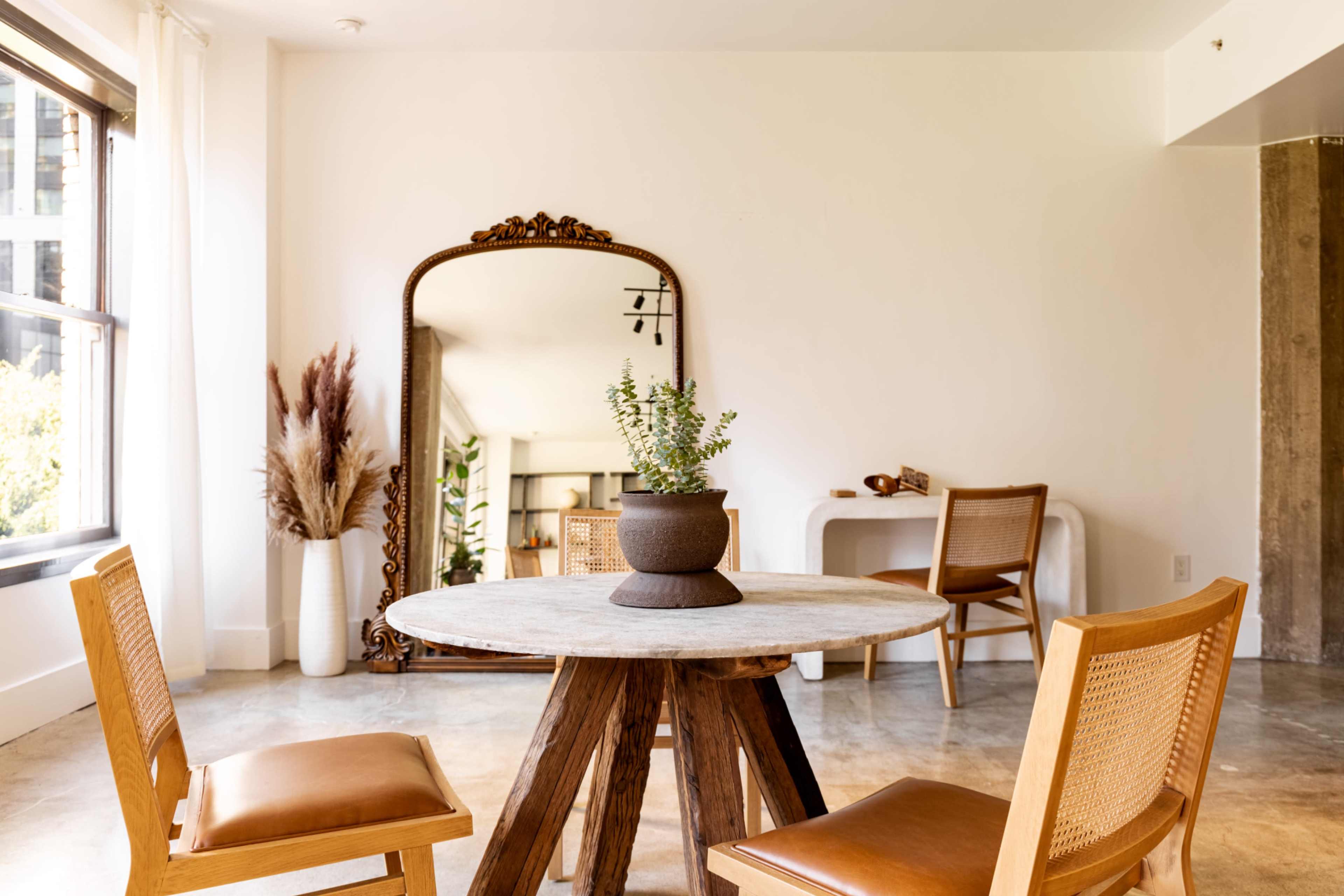 A round wooden dining table with two chairs is placed in a minimalist room featuring a large mirror and a small plant in a pot at the center of the table.