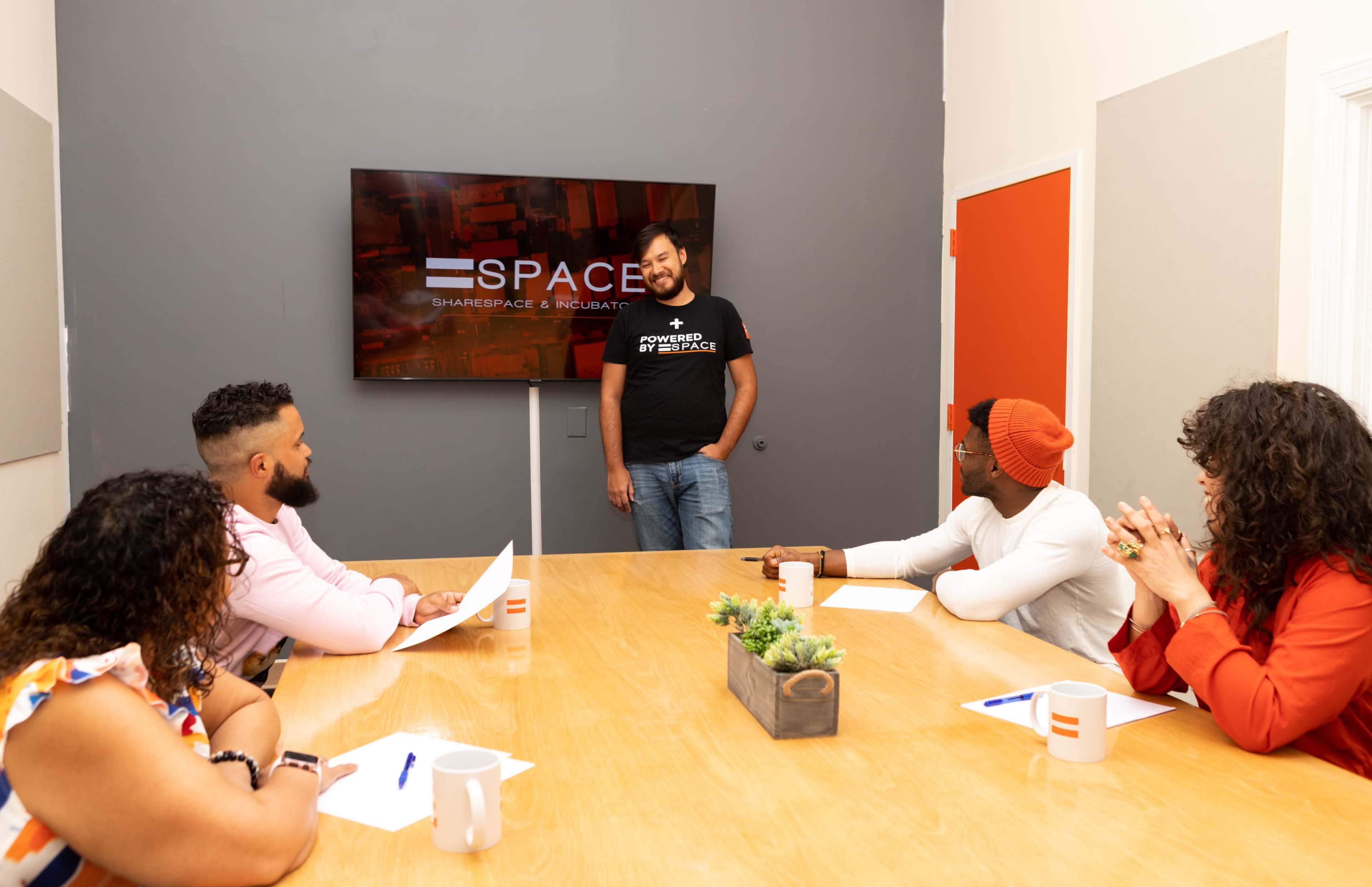 A man stands in front of a television screen displaying "SPACE" while four other individuals sit around a table with notes in front of them.