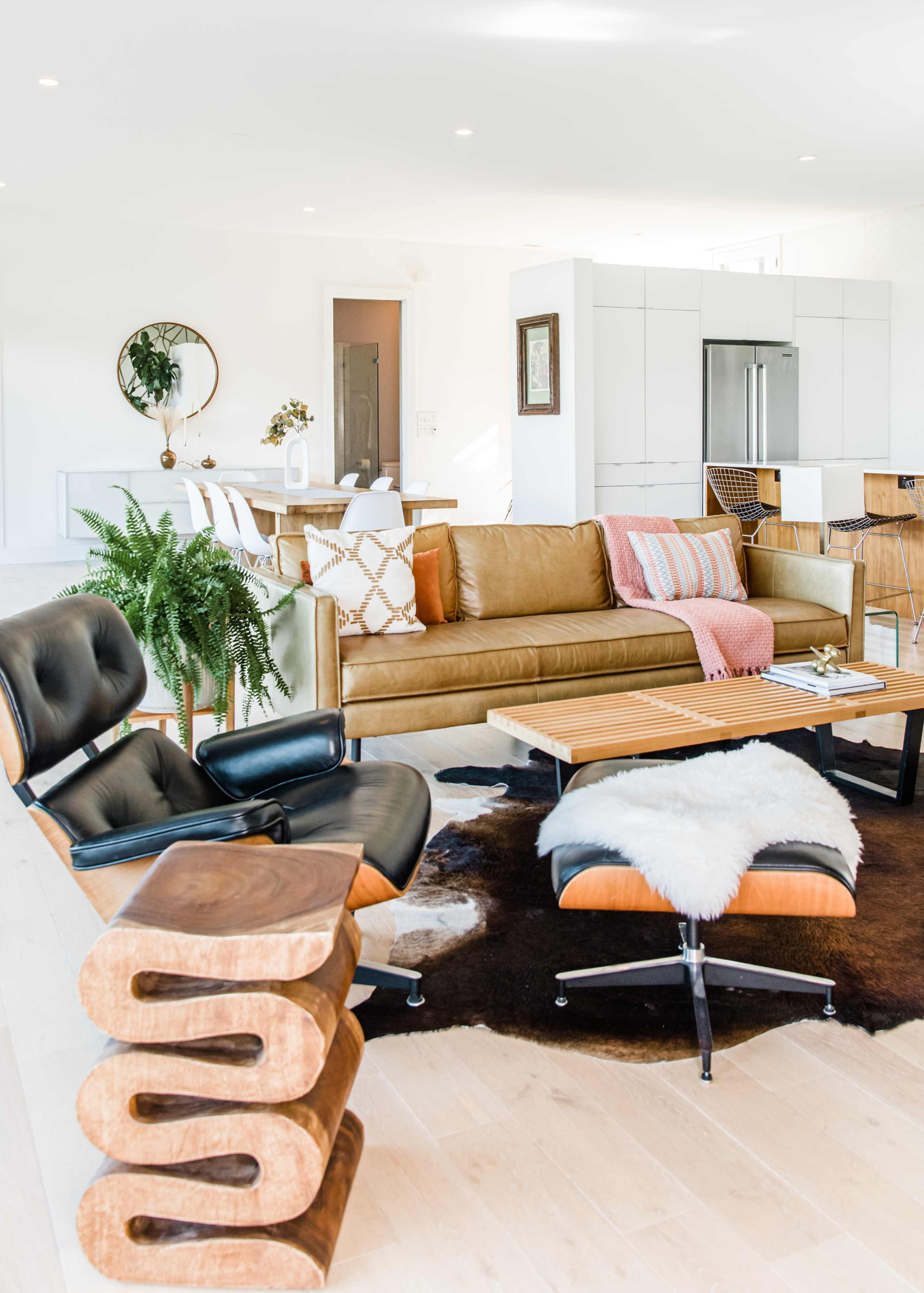 The image shows a modern living room featuring a brown leather sofa, a wooden coffee table, a black and wood swivel chair, and a small centerpiece on a cowhide rug.