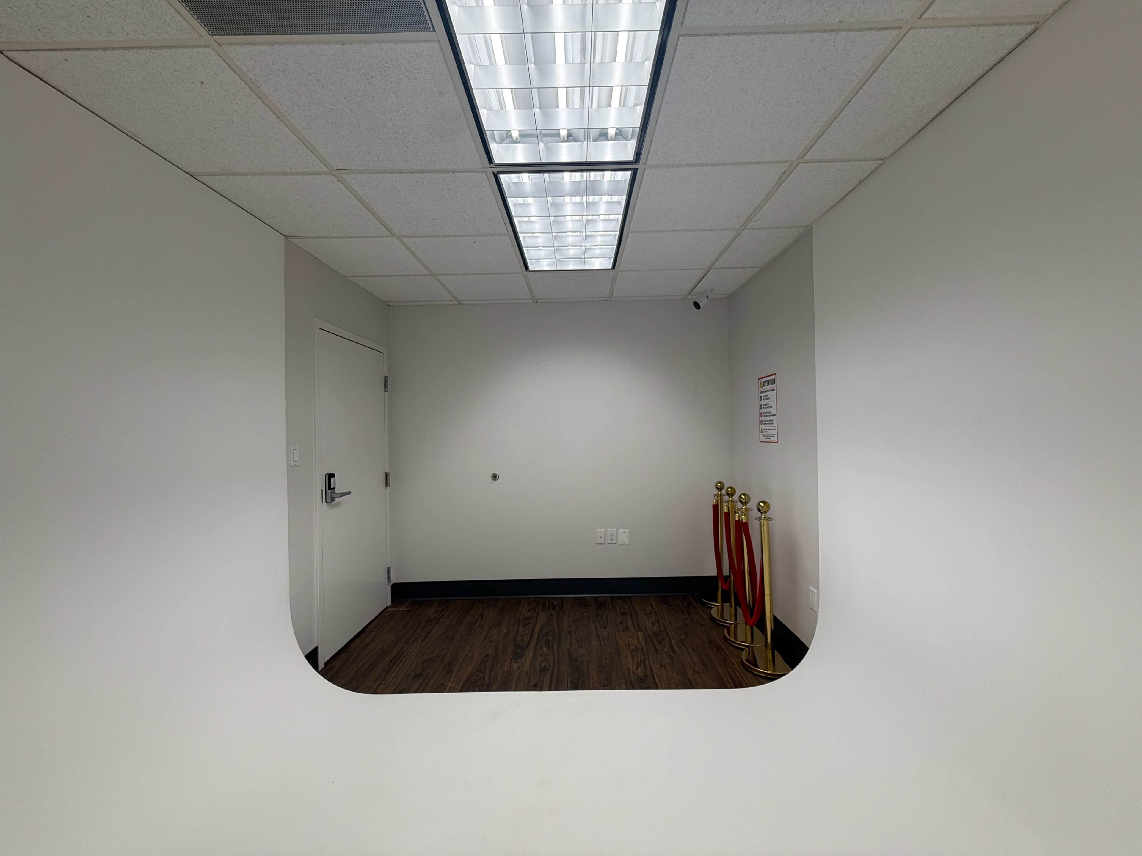 The image shows a small, empty room with an arched opening leading to another space, featuring a wooden floor and two stanchions with red ropes.