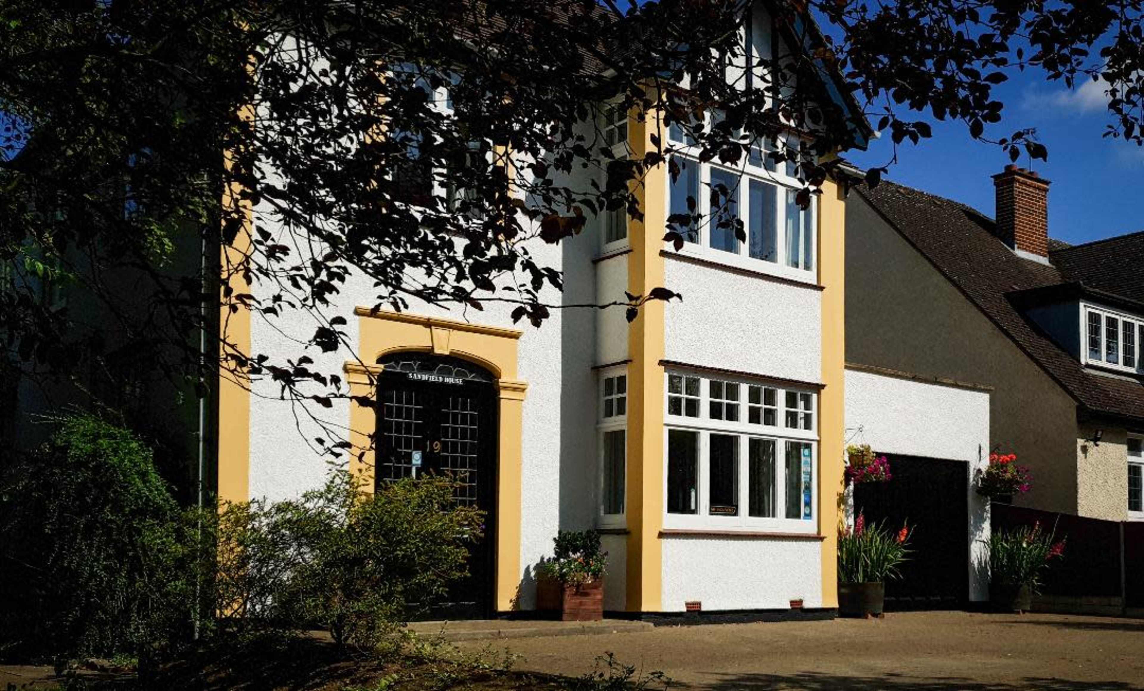 A two-story house with a distinctive yellow trim and large windows is situated in a suburban area, surrounded by trees and flower pots.