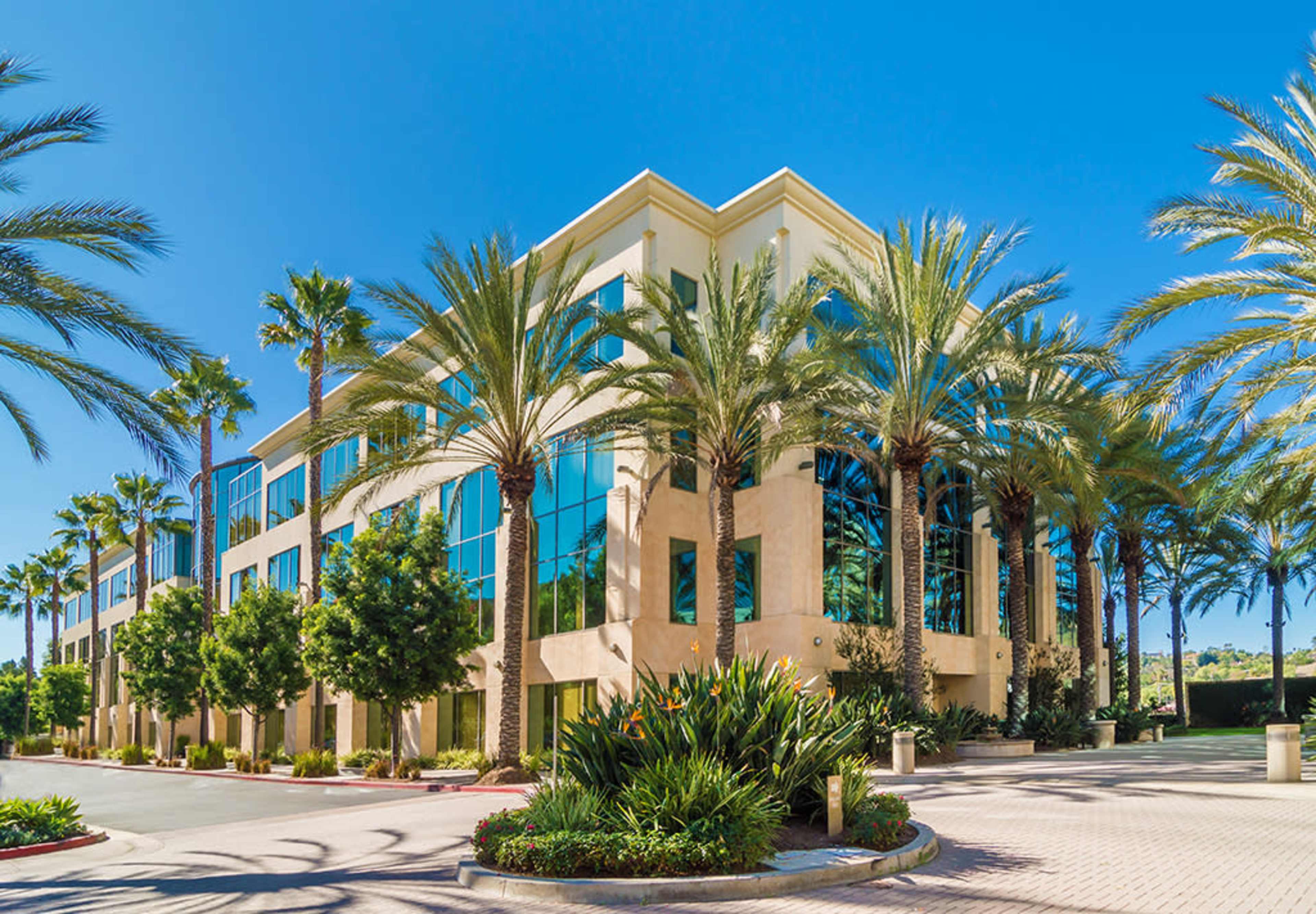 A three-story office building is surrounded by palm trees and landscaped greenery under a clear blue sky.