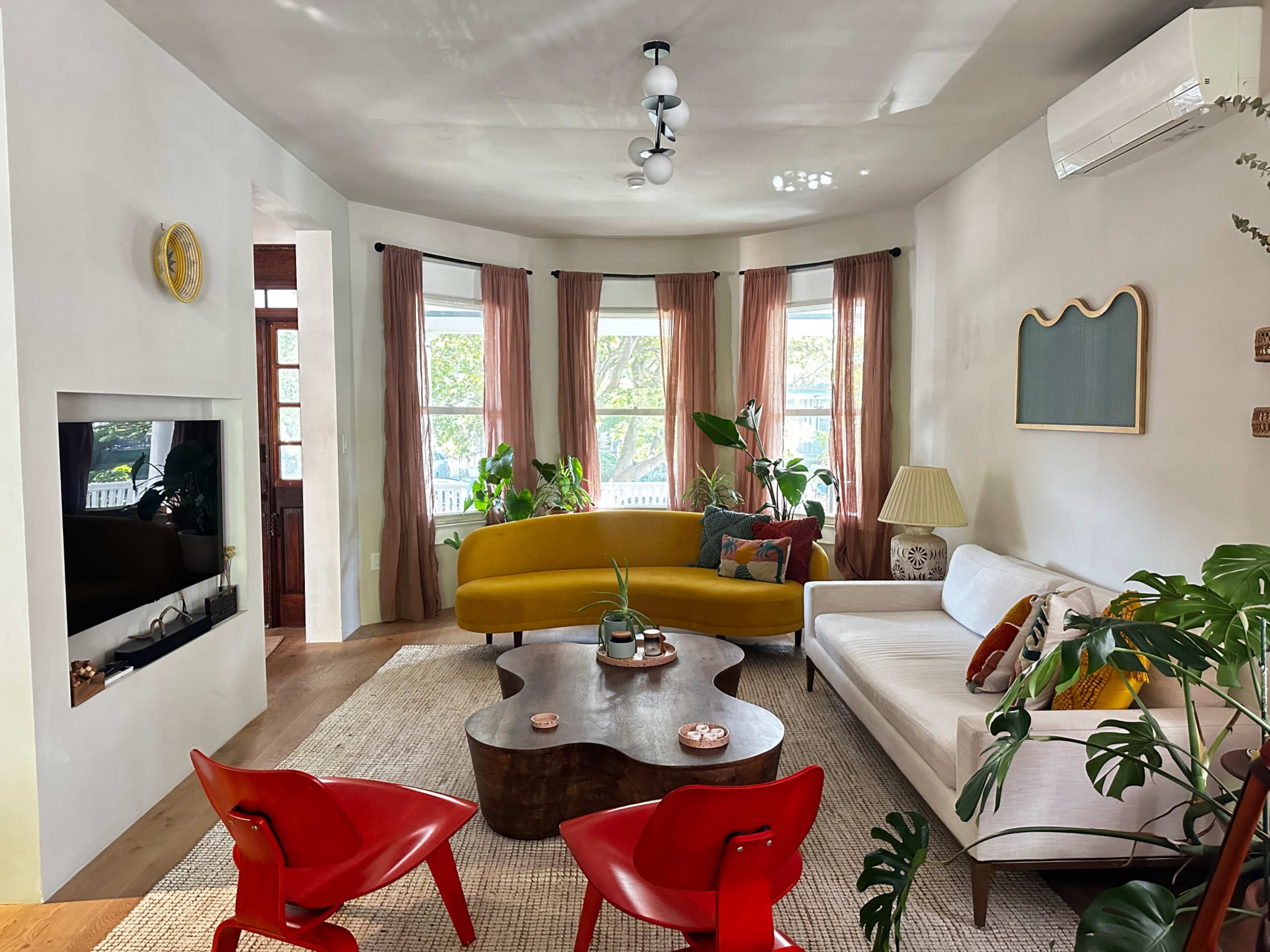 The living room features a curved yellow sofa, a white couch, a round wooden coffee table, and large windows adorned with sheer curtains, surrounded by various plants.