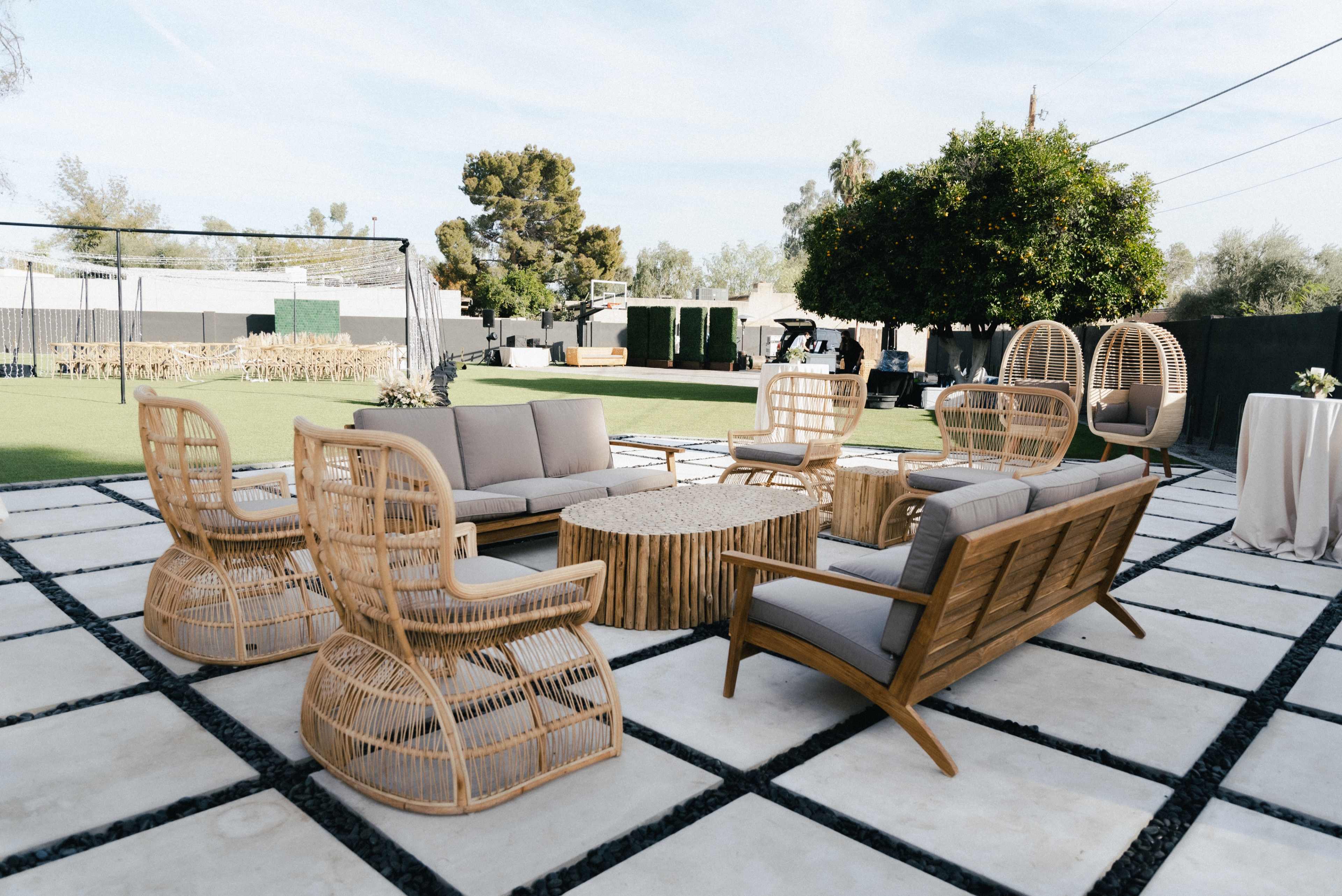 A modern outdoor lounge area featuring woven furniture with a central coffee table, surrounded by a grassy space and trees.