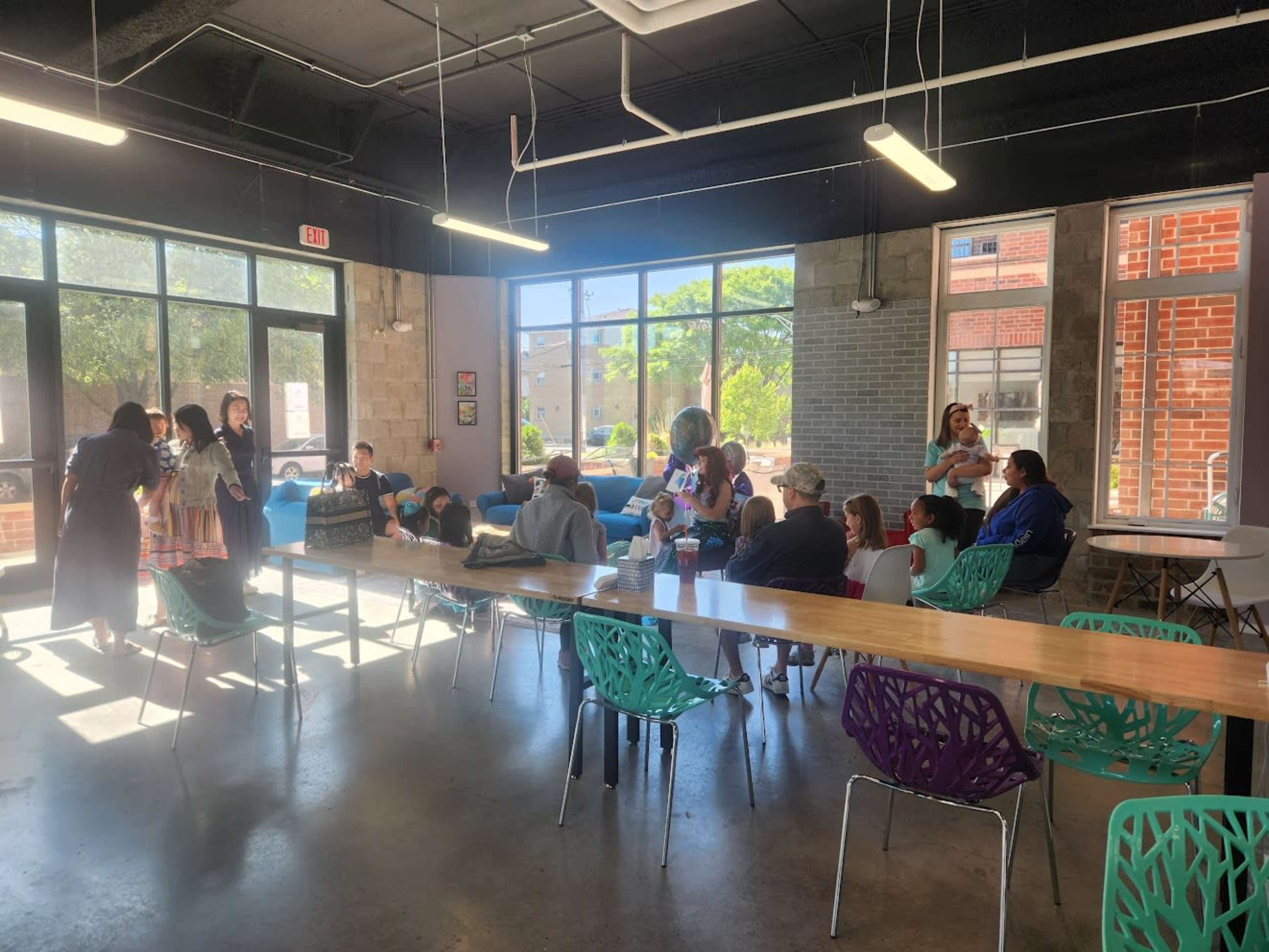 A group of people gathers in a spacious, modern room with large windows and a long table surrounded by colorful chairs.