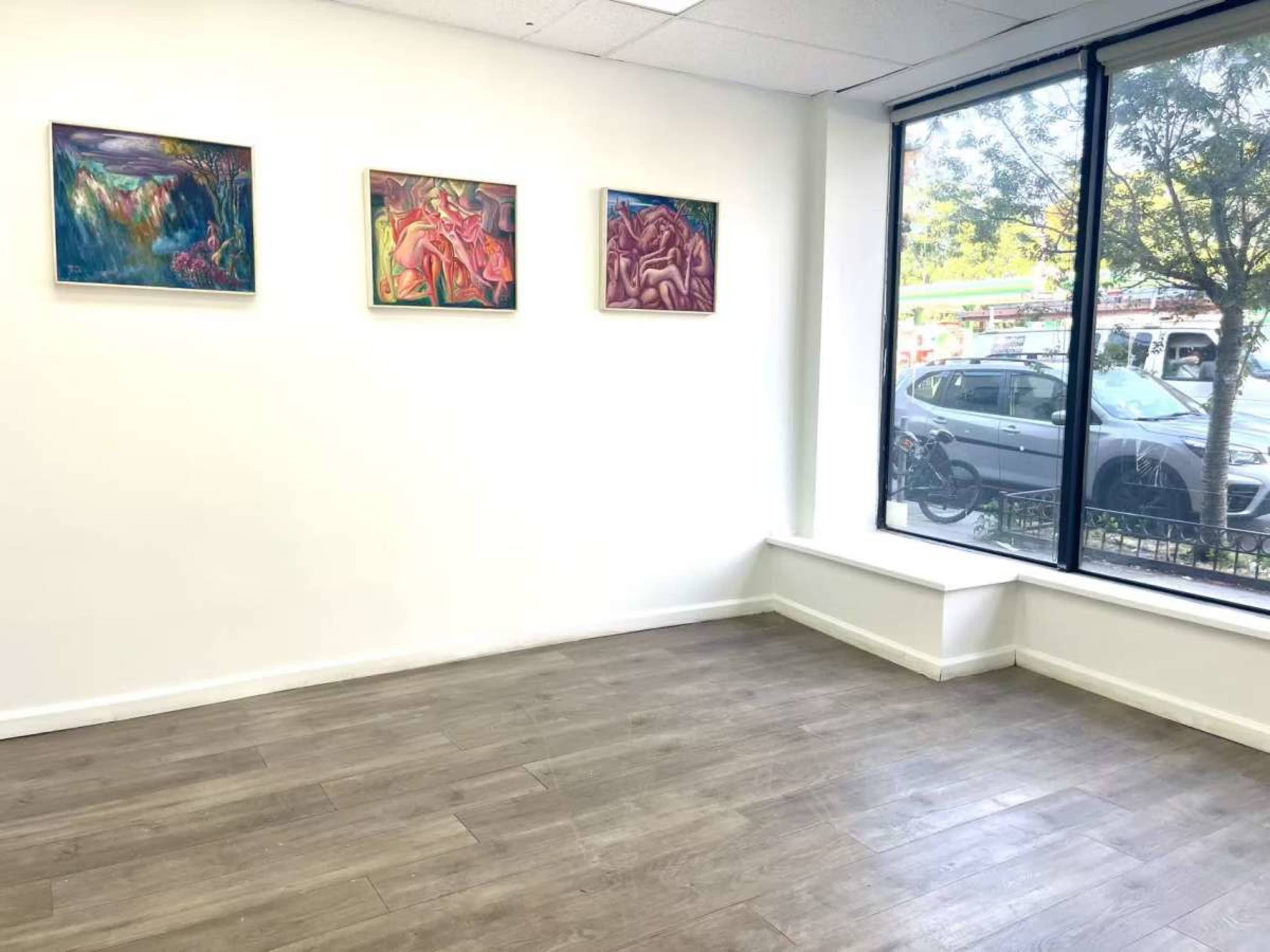 The image shows a bright, empty room with four framed artworks hanging on the wall and a large window providing outside visibility.