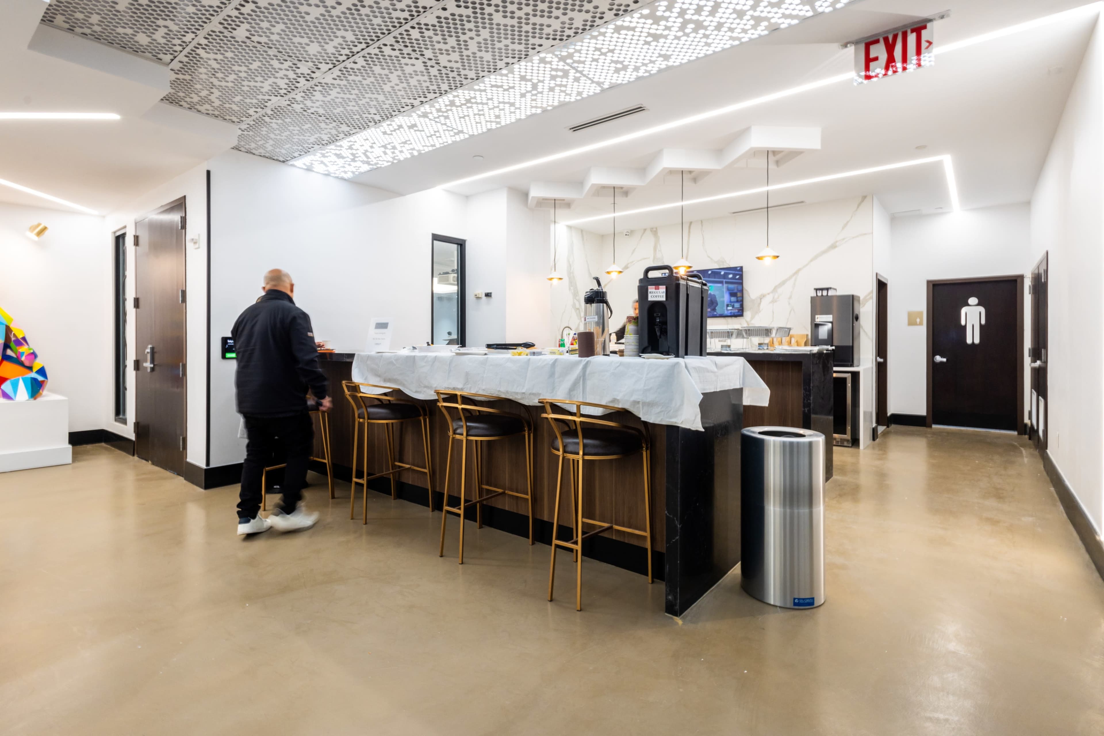 A person walks past a modern kitchen area with bar stools, a coffee machine, and a large wall-mounted TV in a well-lit space.