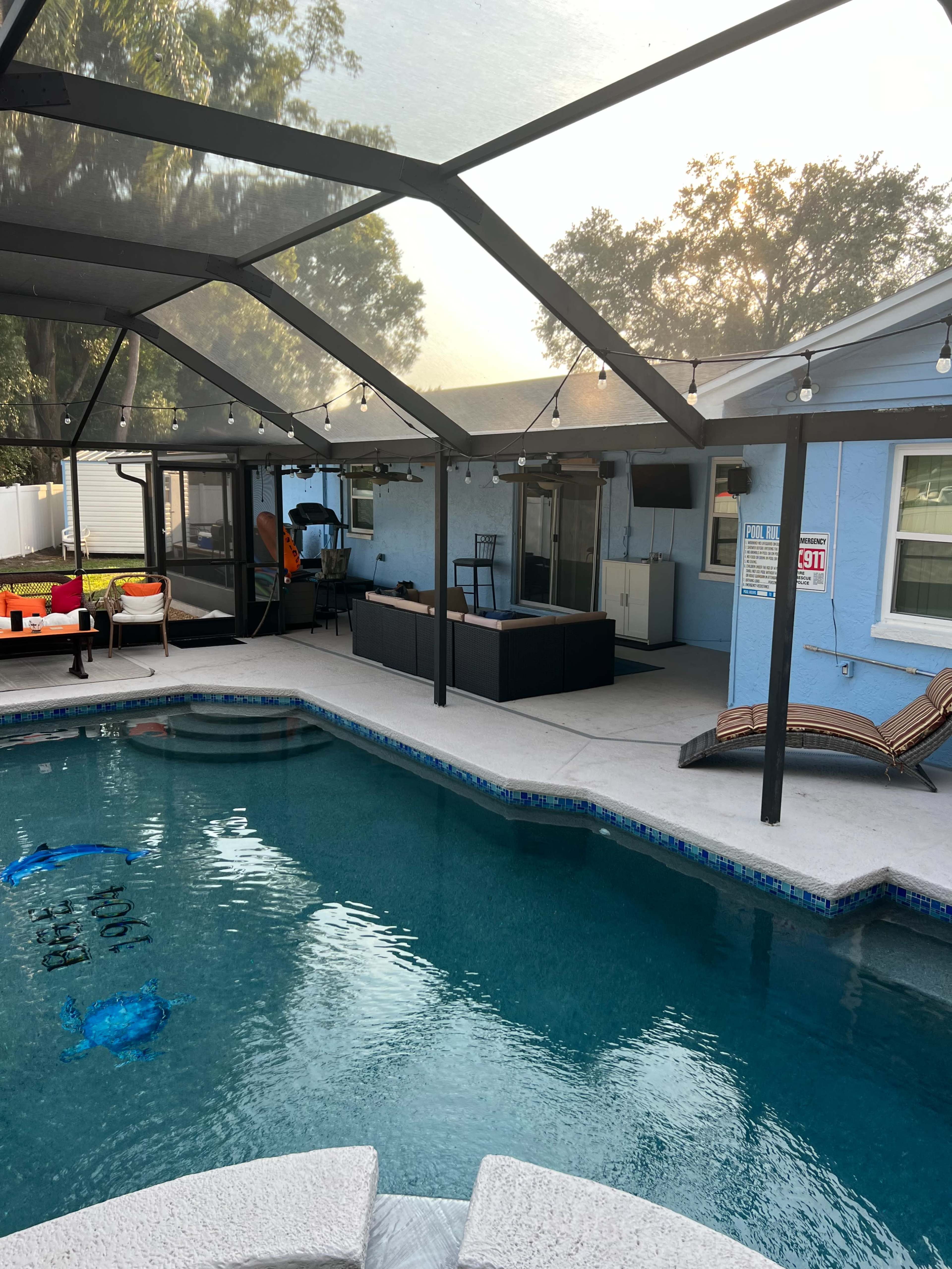 A screened-in outdoor area with a swimming pool, lounge chairs, and a shaded seating space beside a light blue house.