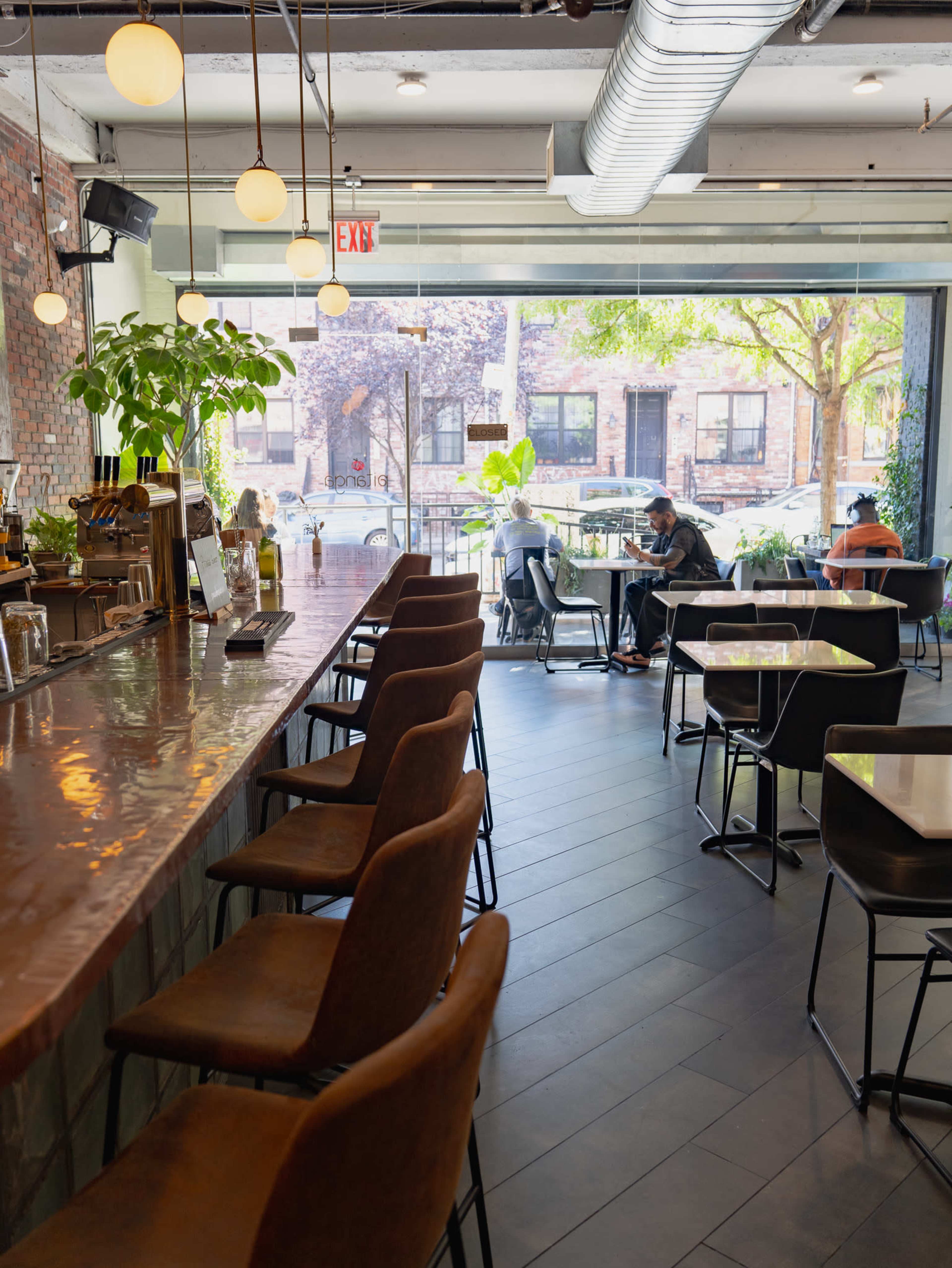 A modern café interior with a long bar on one side, featuring seating for patrons, and tables scattered throughout, with large windows overlooking a tree-lined street.