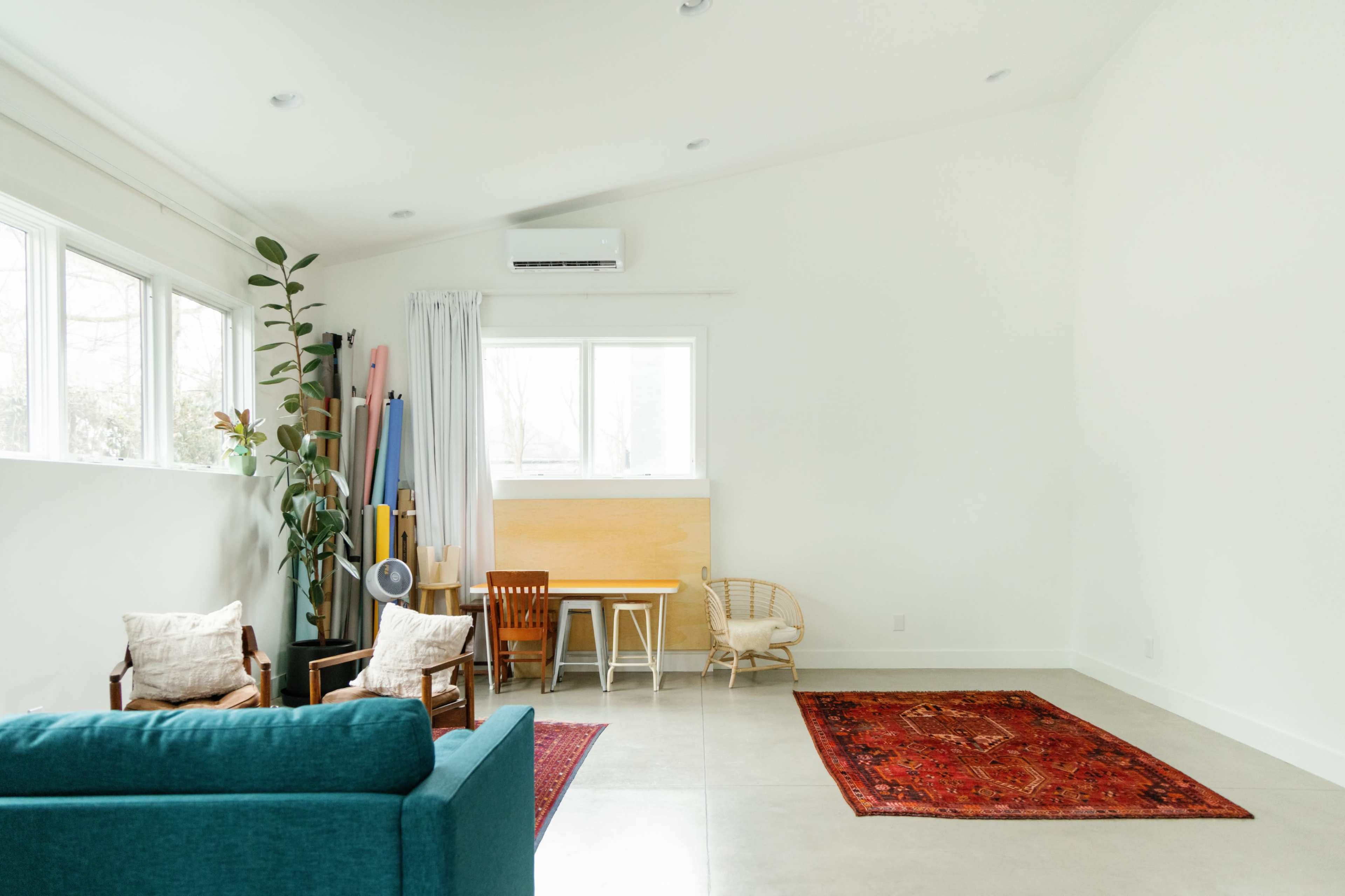 The room features a simple design with a blue couch, chairs, a wooden table, and large windows allowing natural light to fill the space.