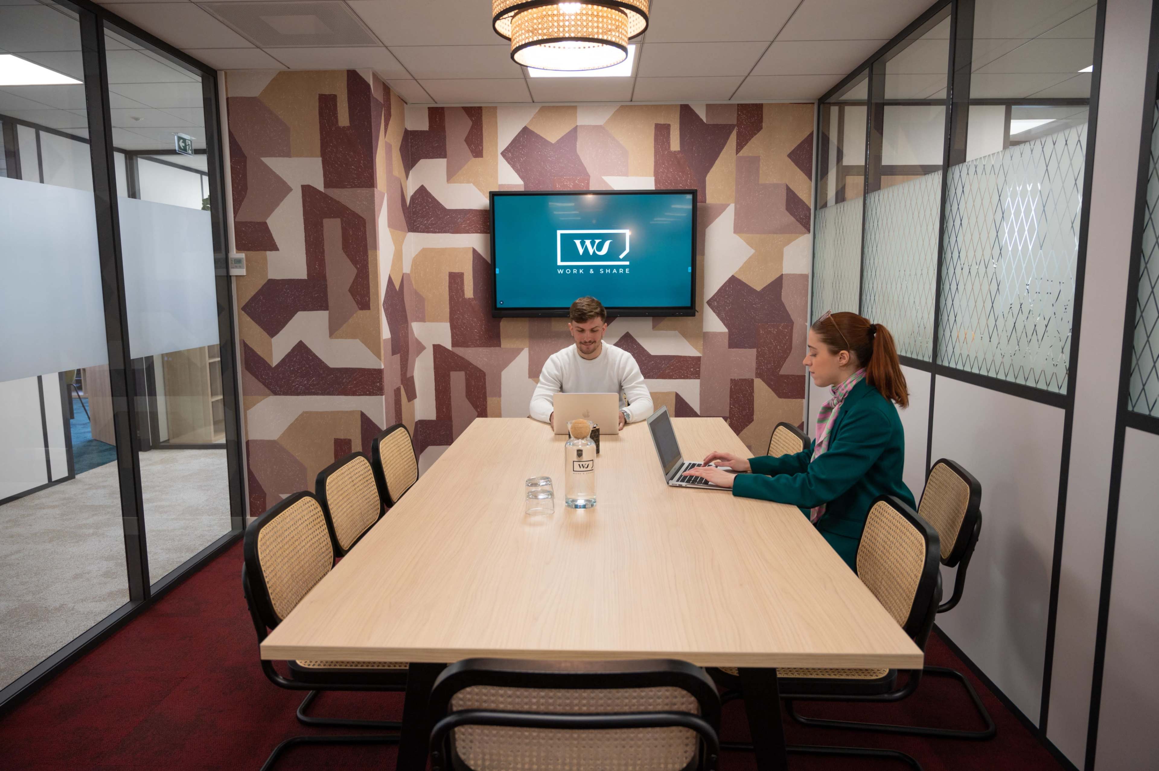 A conference room features a long table with two people seated, one working on a laptop and the other typing on a computer, with a TV displaying a logo in the background.