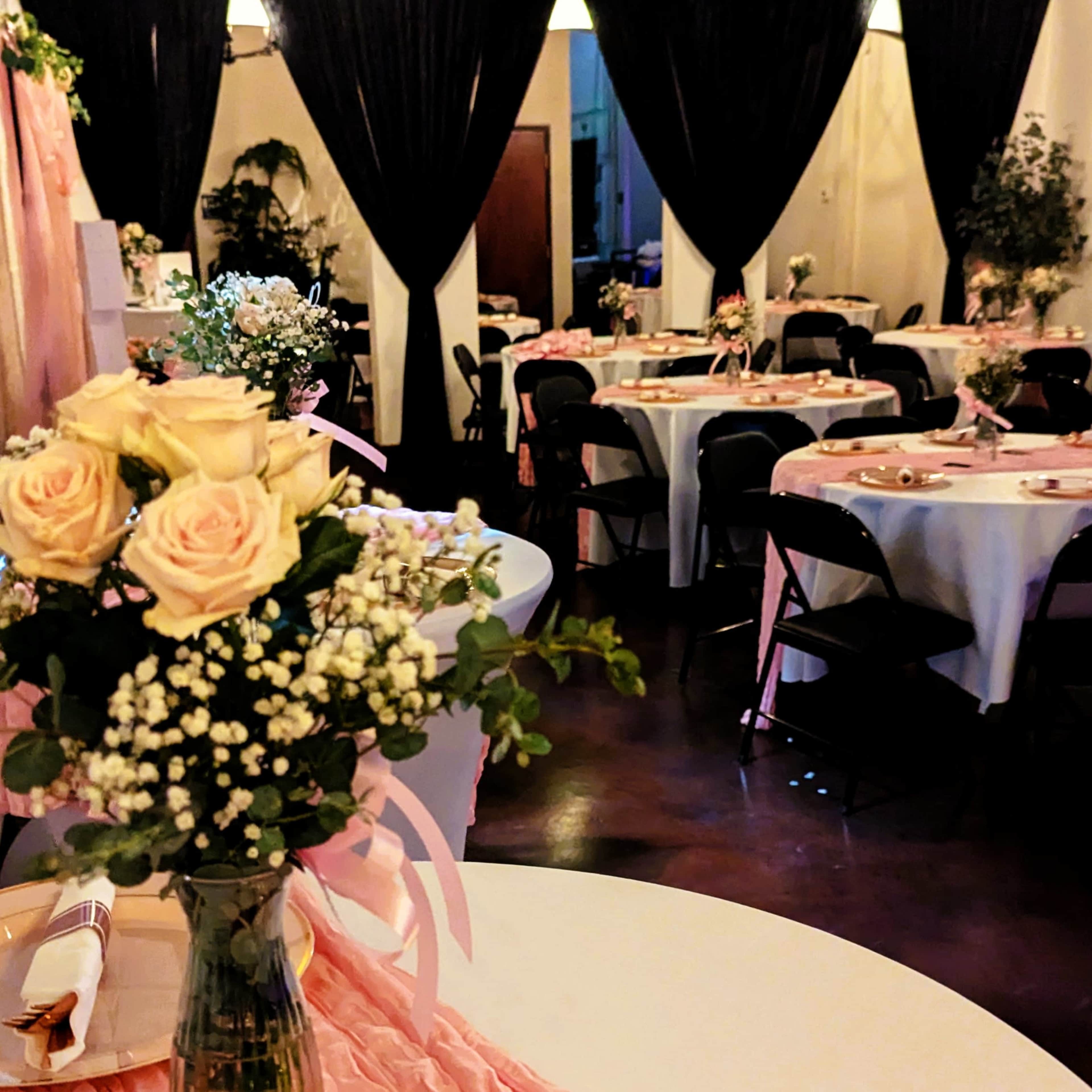 The image shows a decorated event space with round tables set for dining, adorned with flowers and draped fabric in the background.