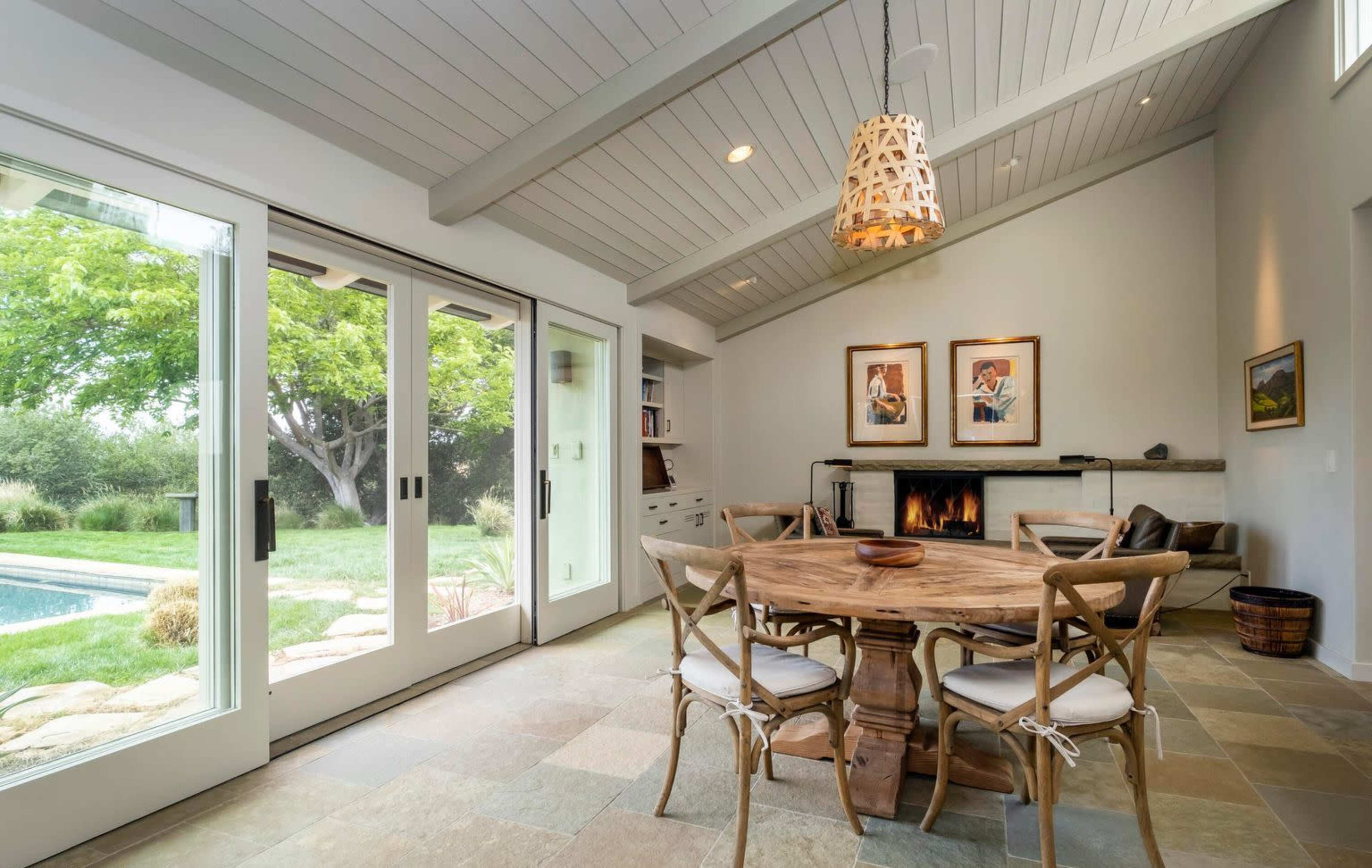 A modern dining area with a wooden table surrounded by chairs, large glass doors opening to an outdoor space, and a fireplace along one wall.