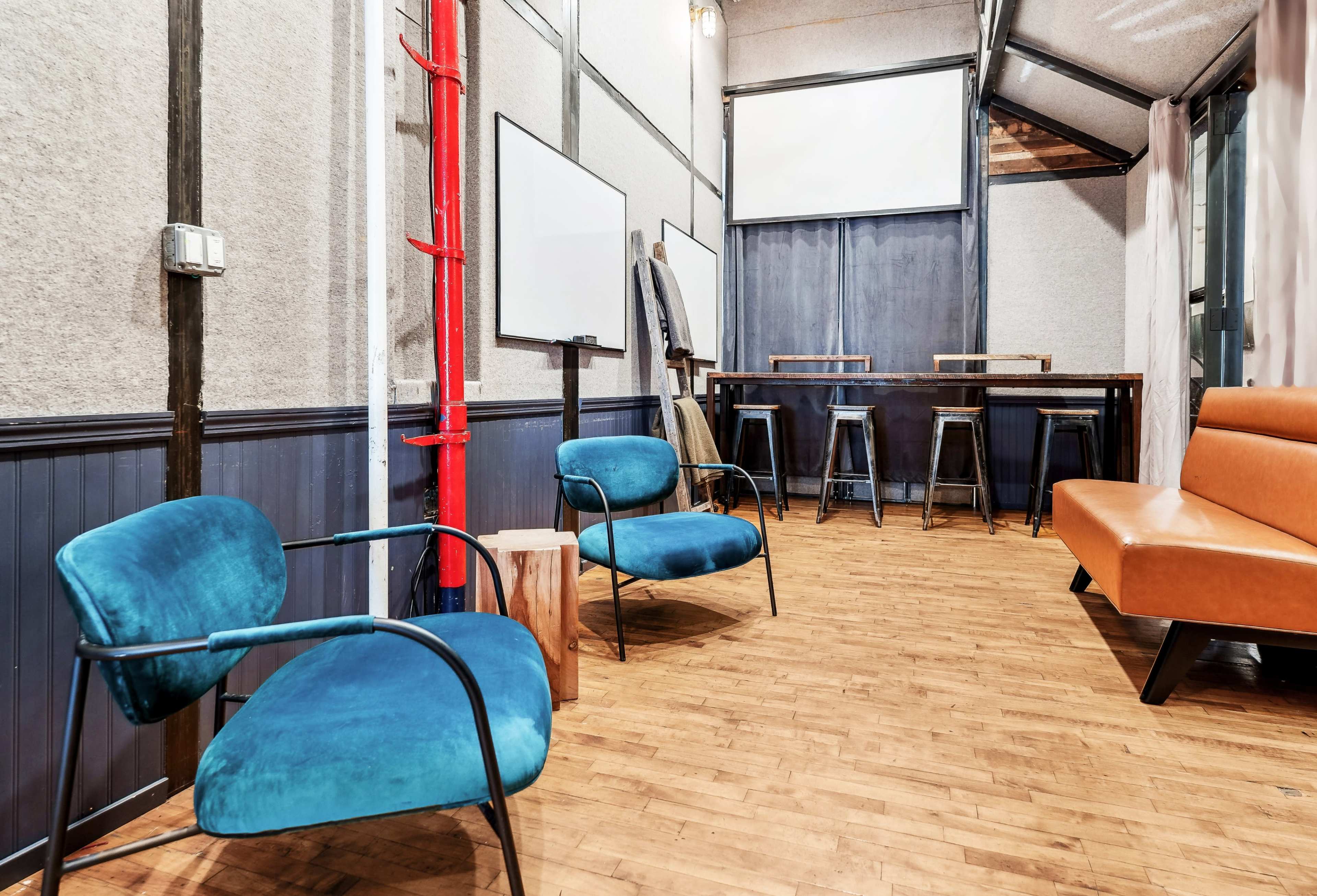 The image shows a modern interior lounge area with blue chairs, wooden flooring, and minimalist decor, including tables and wall screens.