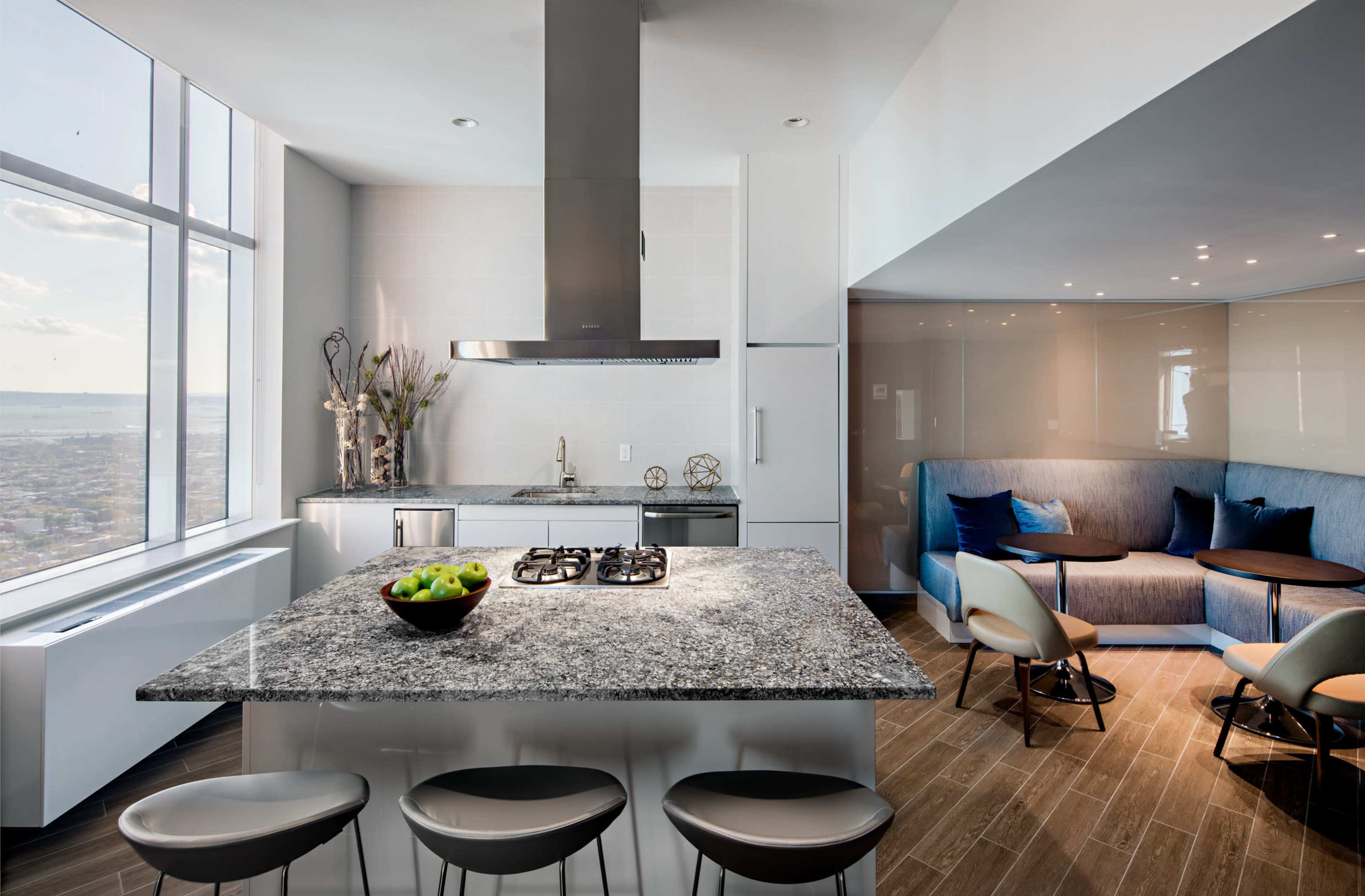 The image shows a modern kitchen featuring a large granite island with a gas cooktop, sleek cabinetry, and a seating area with curved chairs and a sofa, all illuminated by natural light from large windows.