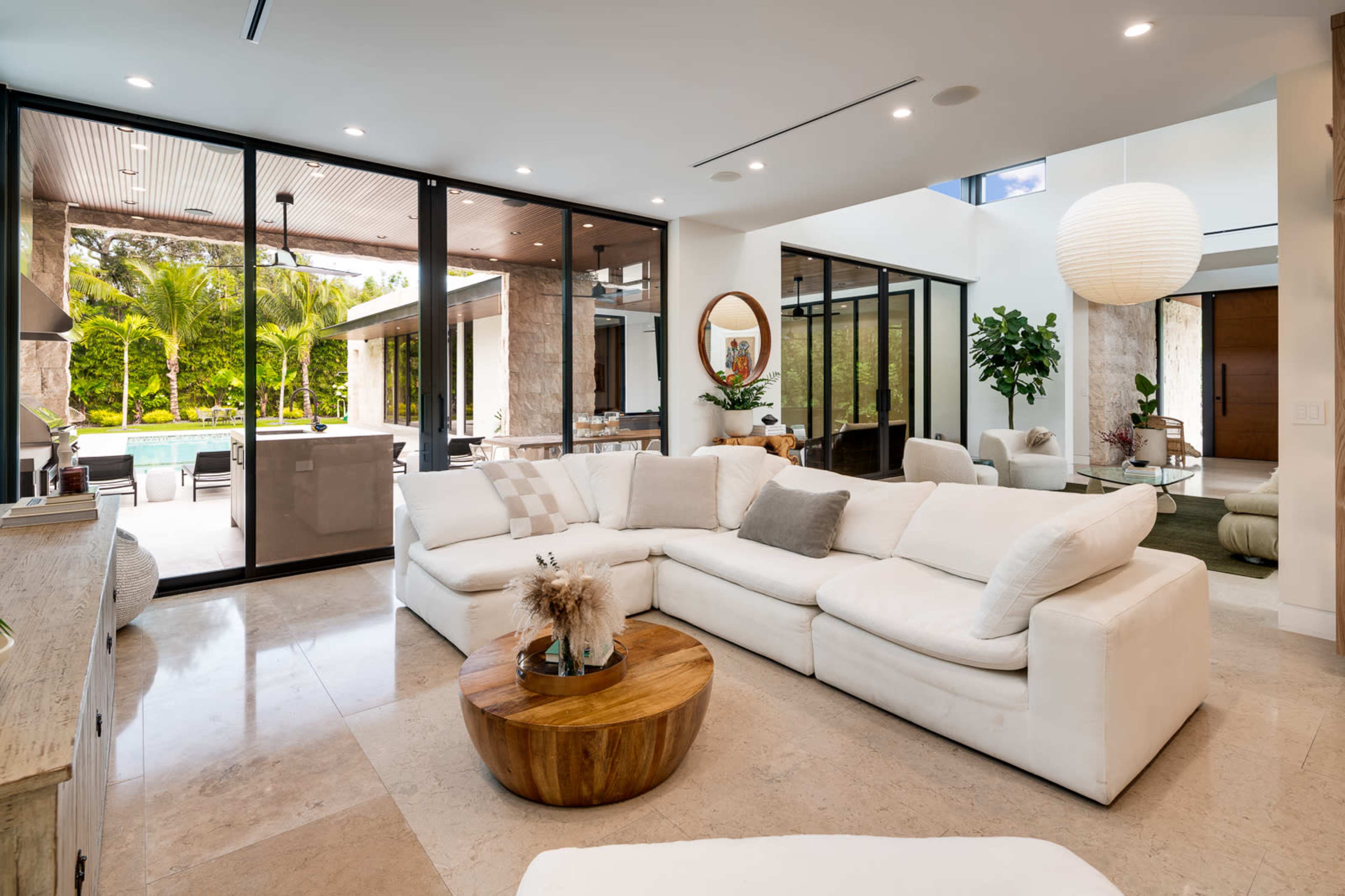 The image shows a modern living room featuring a large white sectional sofa, a round wooden coffee table, and floor-to-ceiling glass doors that open to a patio with greenery.