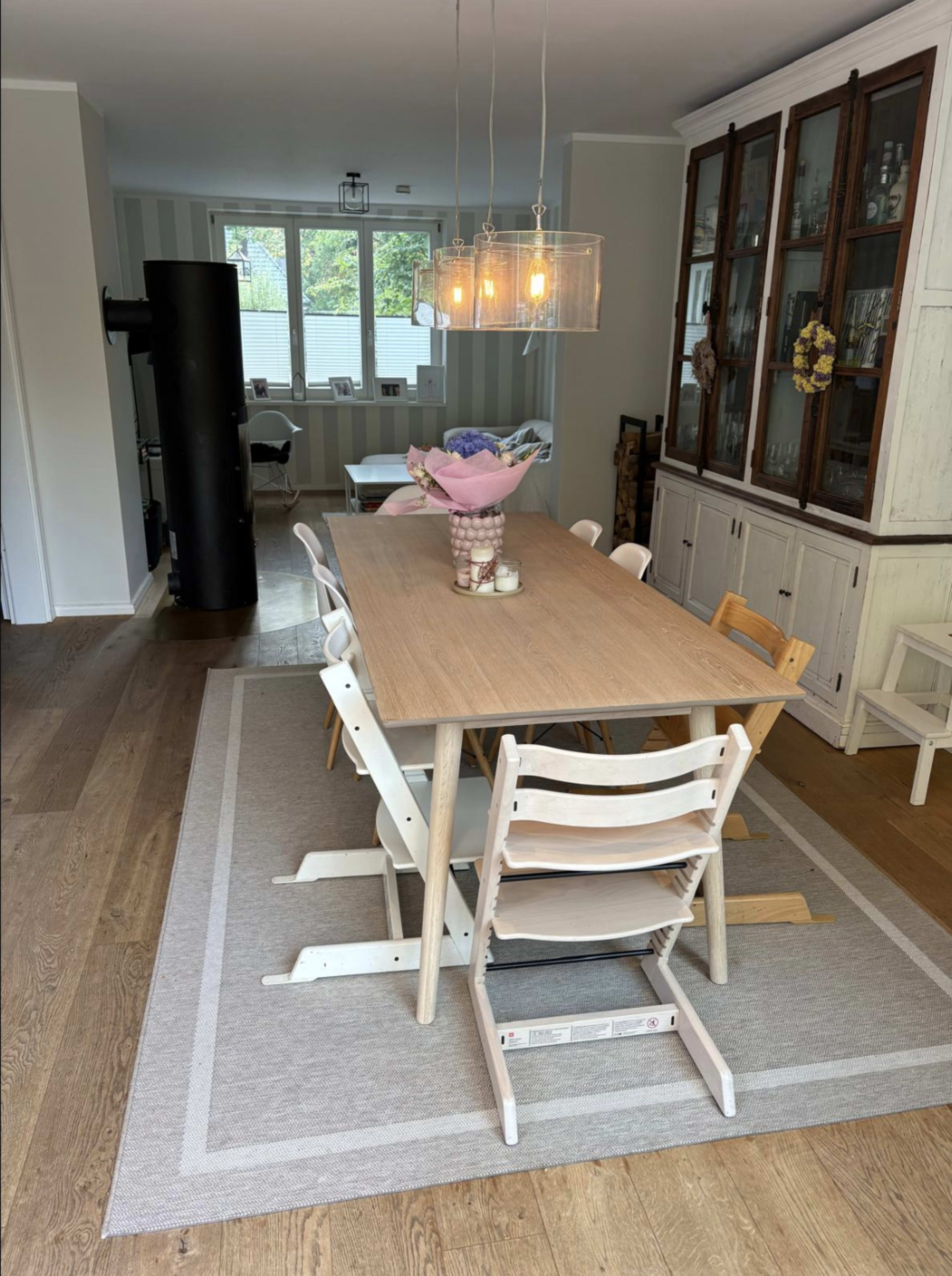 A wooden dining table surrounded by chairs is set in a well-lit dining area featuring a light rug and a glass-front cabinet.