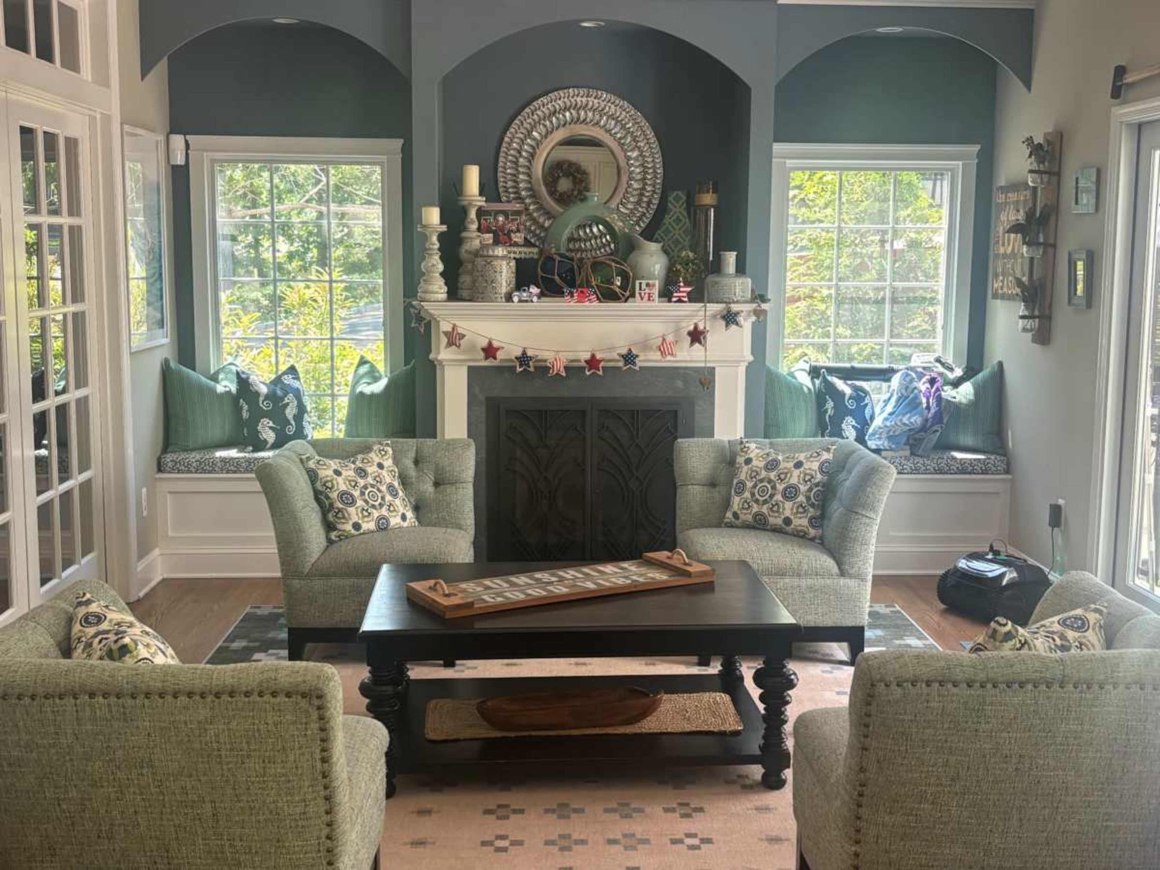 The image shows a cozy living room with two armchairs facing a coffee table, a fireplace adorned with decor, and a window seat lined with cushions.