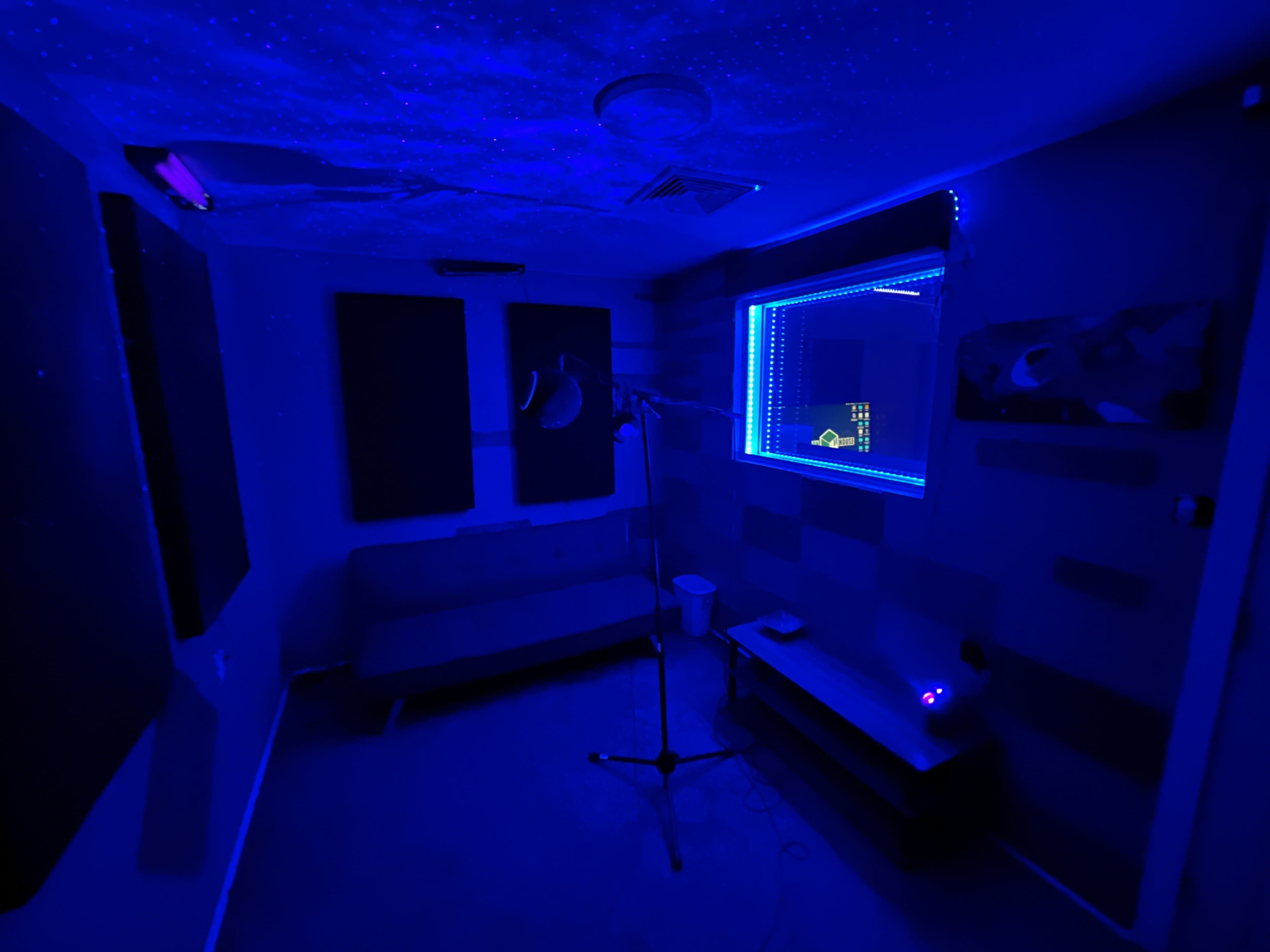 A small, dimly lit recording studio featuring a microphone on a stand, a couch, and a window framed with blue LED lights.