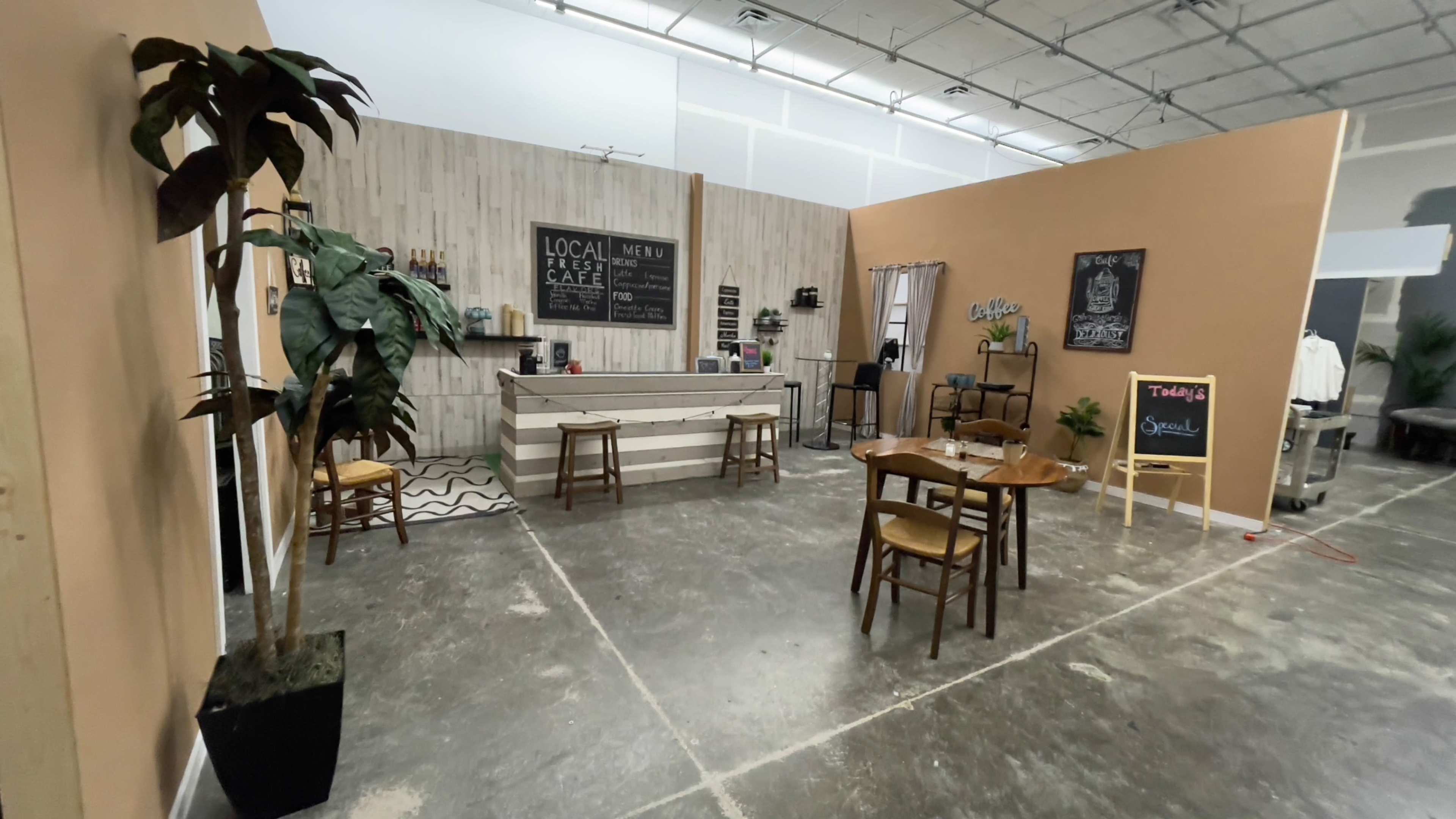 The image shows a modern café interior with a wooden counter, chairs, tables, and décor against a neutral backdrop.
