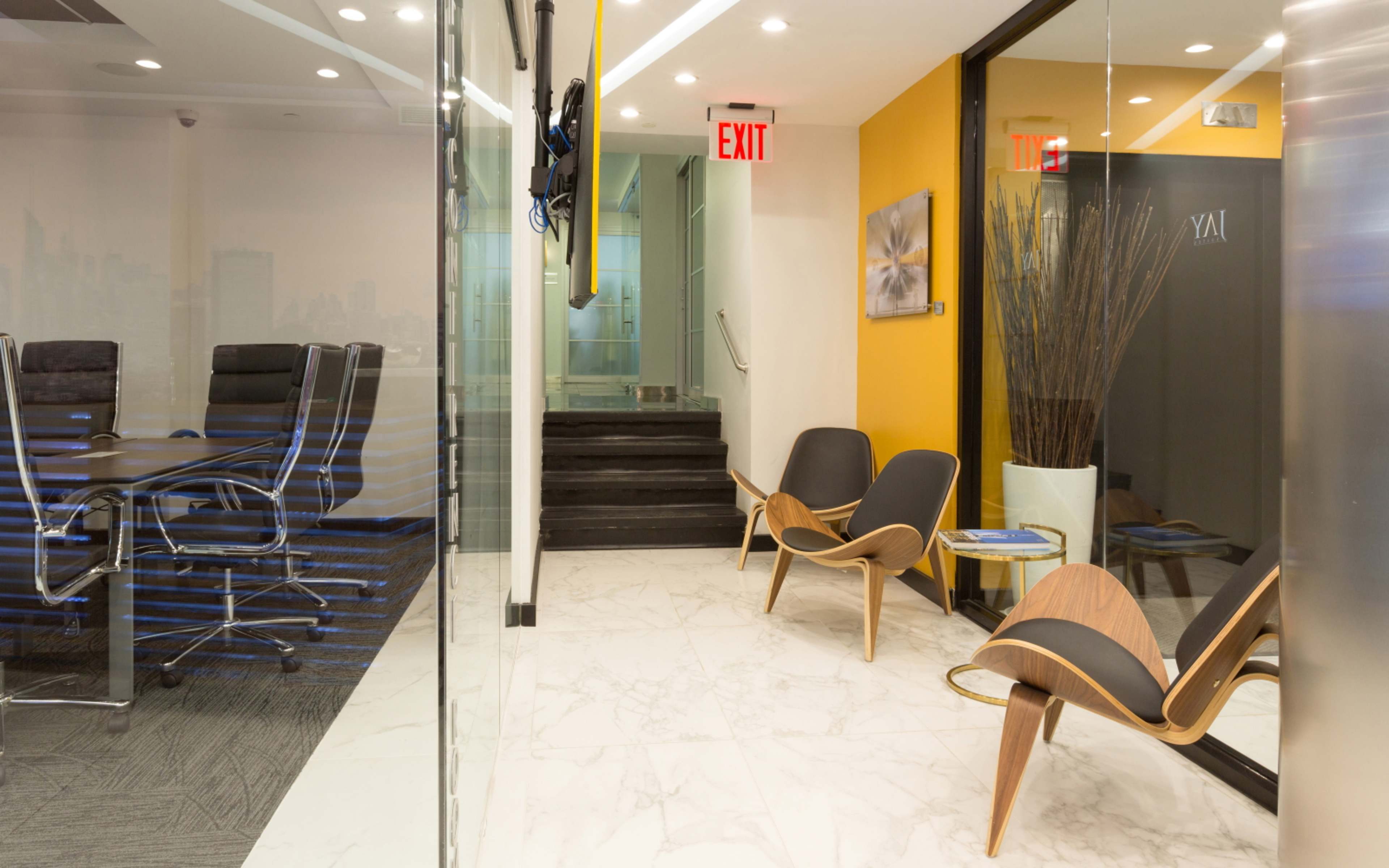 A modern office space featuring a glass meeting room with a conference table and chairs, alongside a foyer area with stylish seating and yellow accent walls.