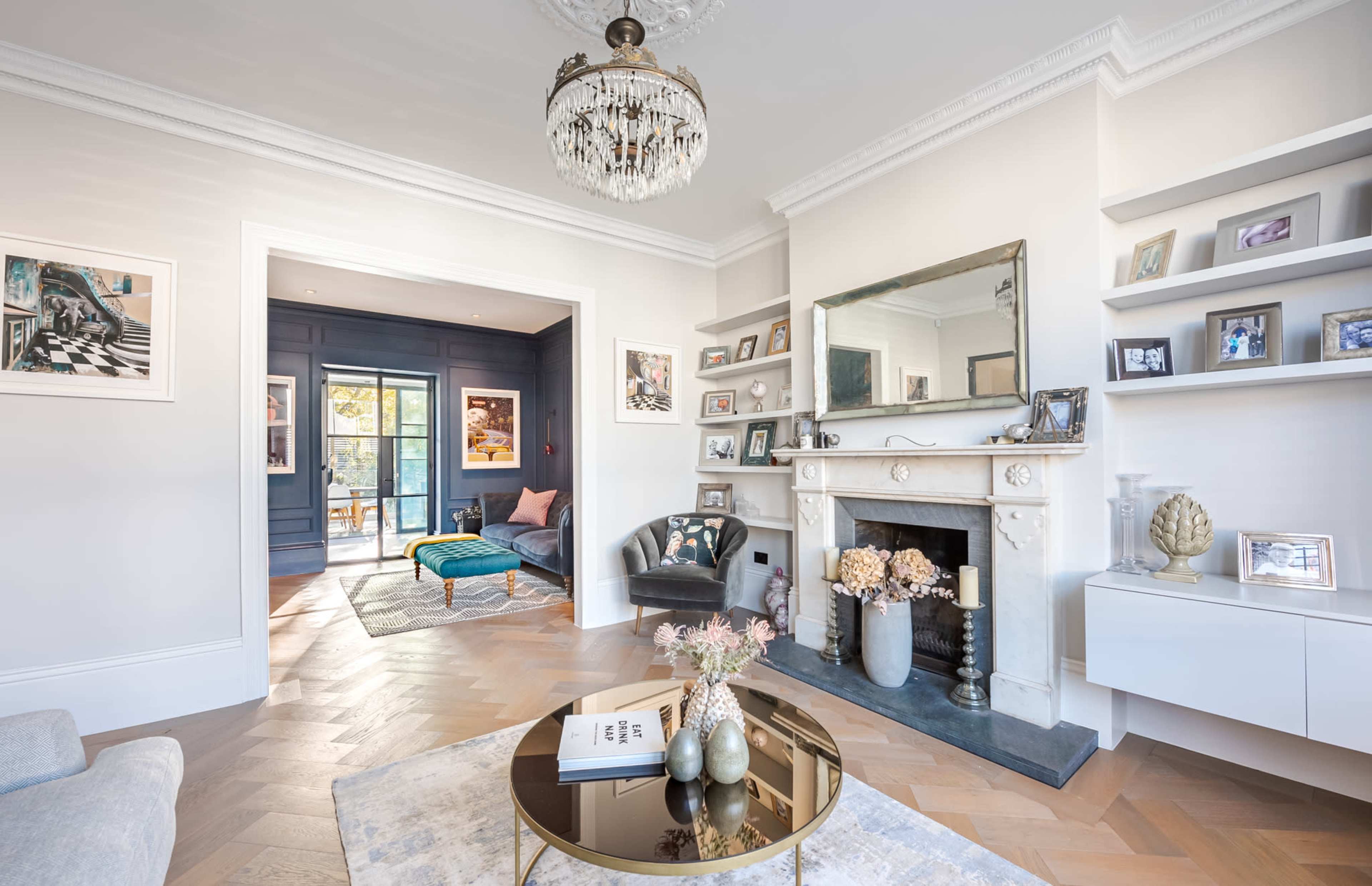 The living room features a chandelier above a coffee table, a fireplace with decorative elements, and access to a sunlit area through large glass doors.