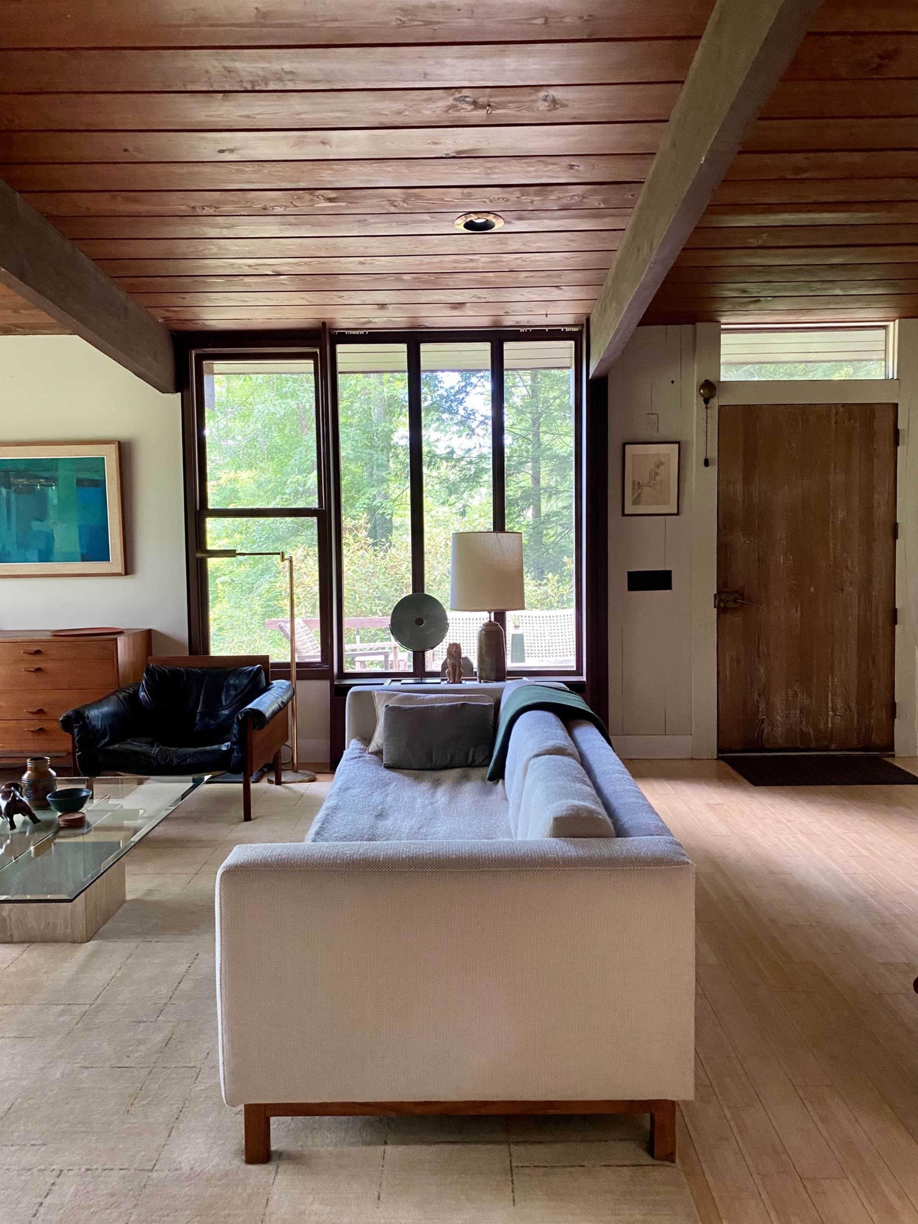 The image shows a modern living room with a sofa, coffee table, and large windows overlooking greenery.