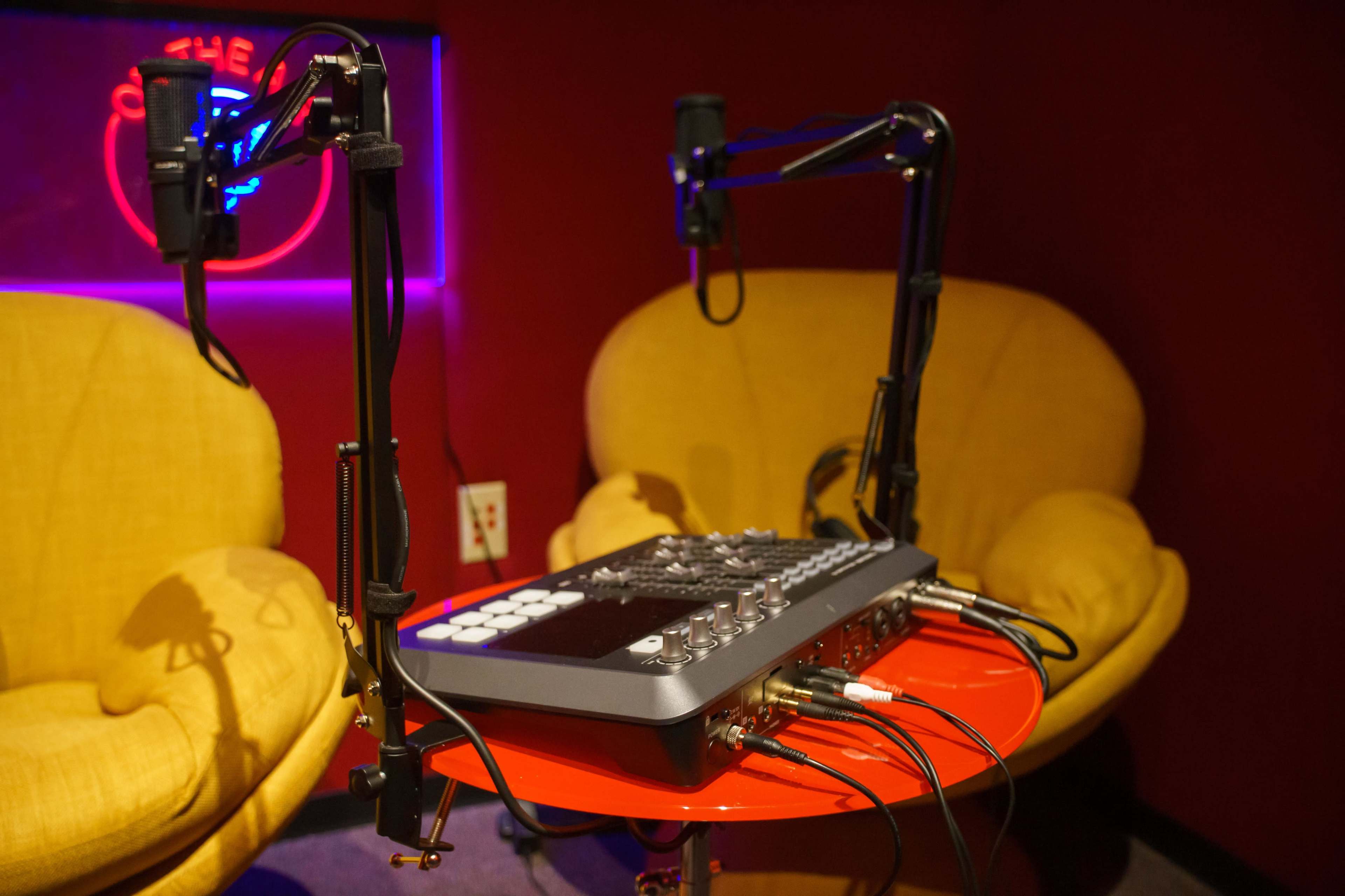 A podcasting setup featuring a mixing console with microphones on adjustable stands beside two yellow chairs against a red wall with neon lighting.