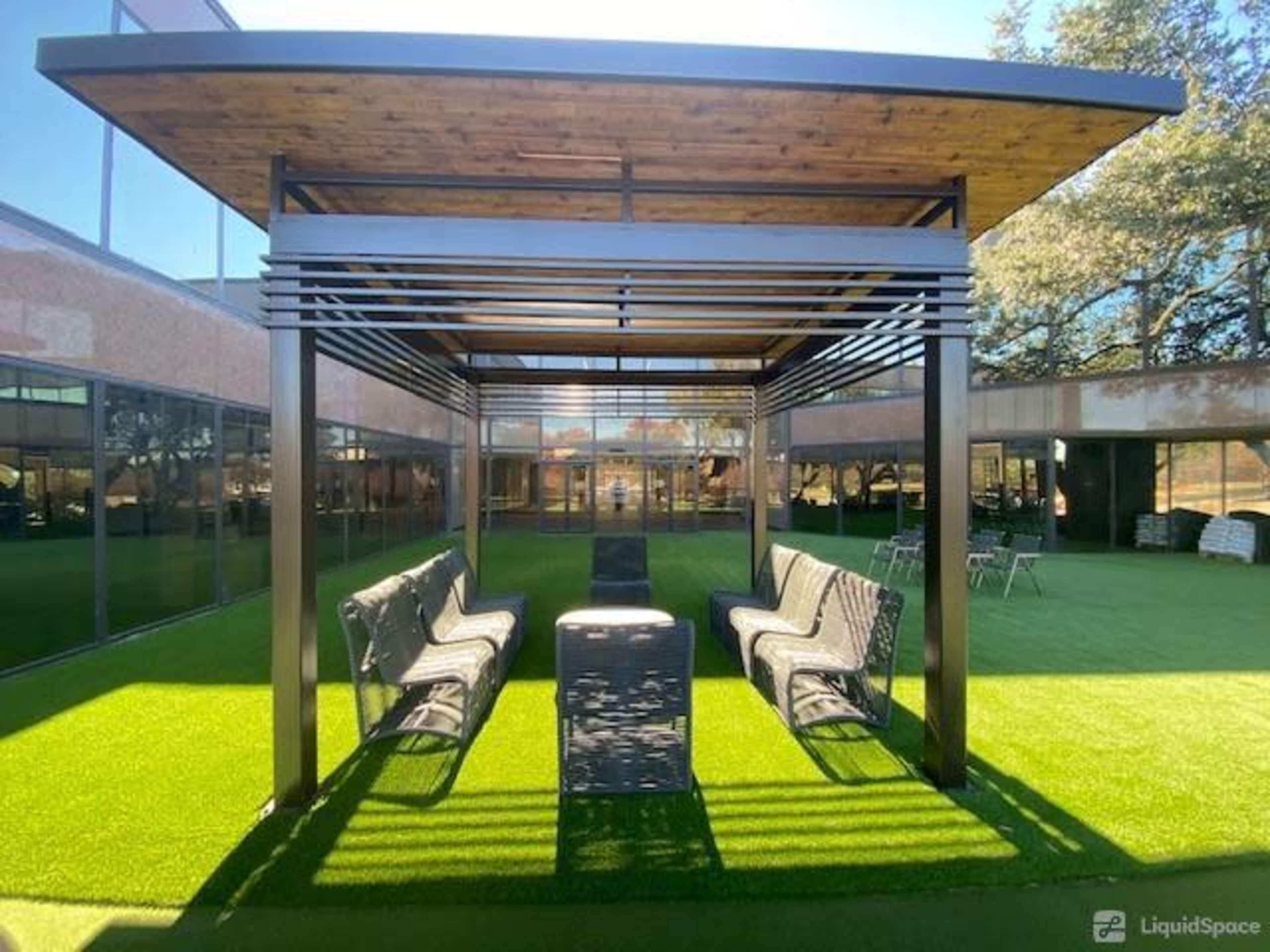 A shaded seating area with modern benches is situated on artificial turf, framed by a building with large windows.