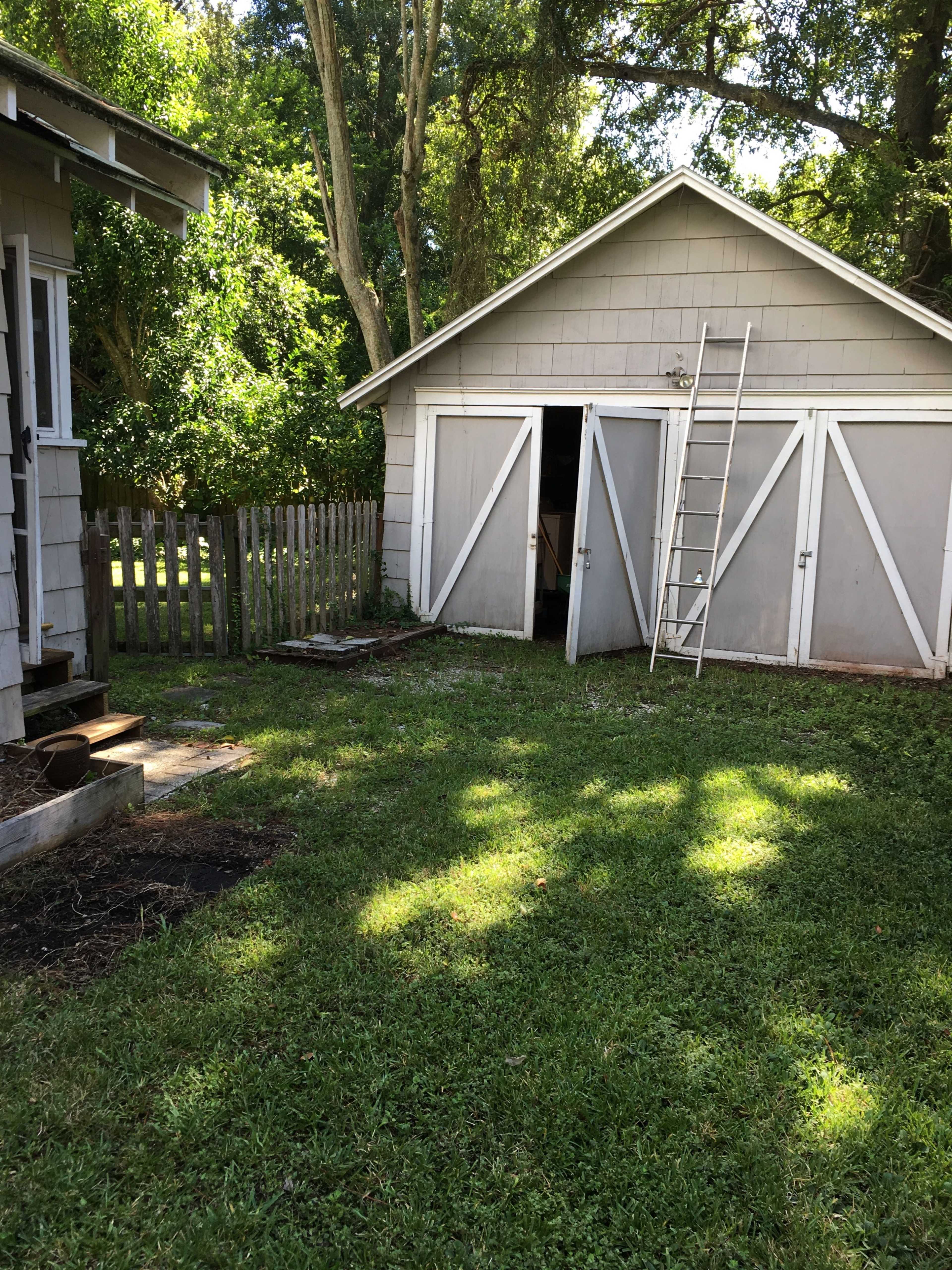 A gray shed with a ladder leaning against it is situated in a grassy area next to a house and a wooden fence.