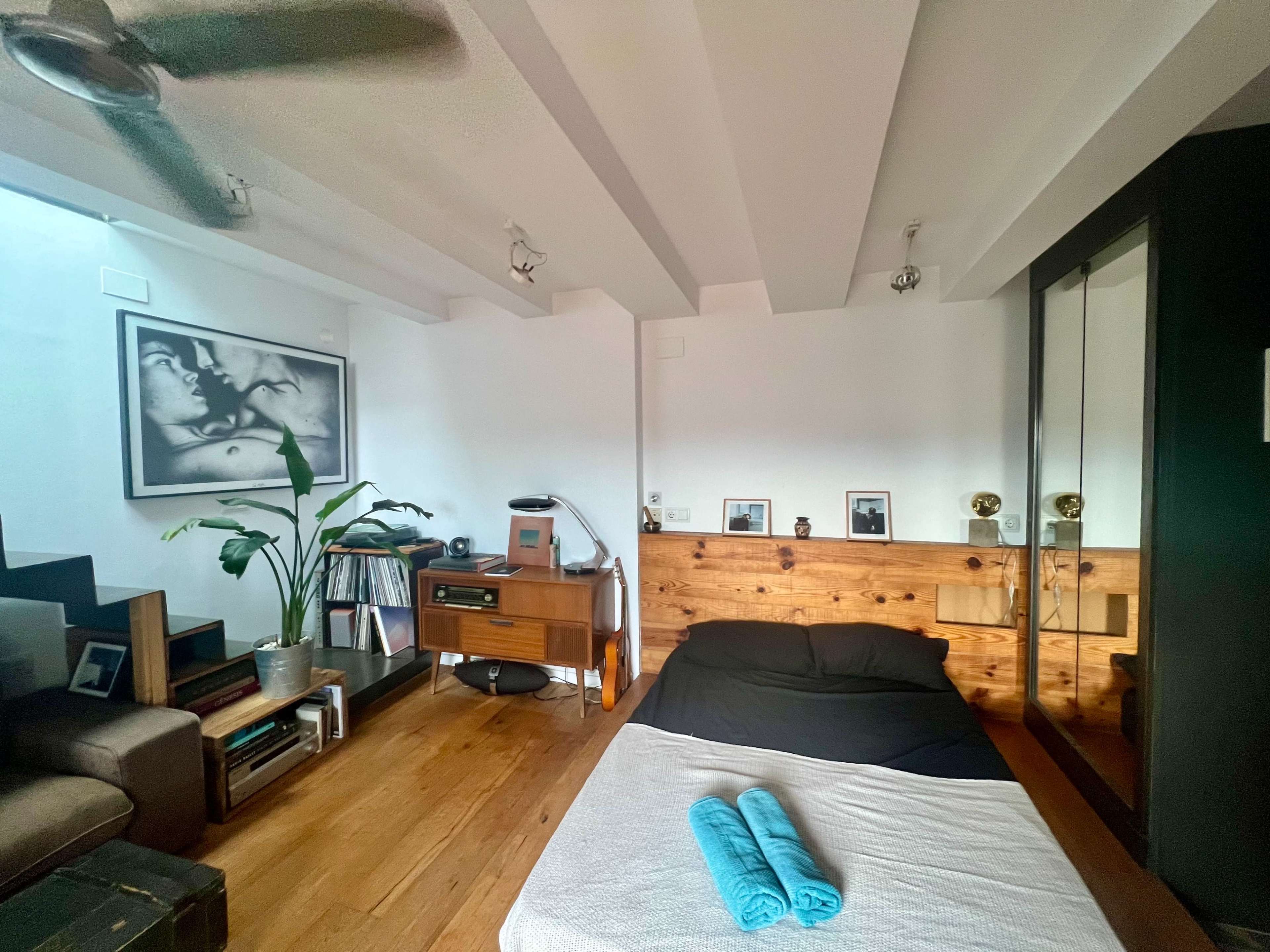 The image shows a modern, minimalist bedroom featuring a wooden floor, a black bed with folded turquoise towels, a sectional sofa, a staircase, and framed artwork on the walls.