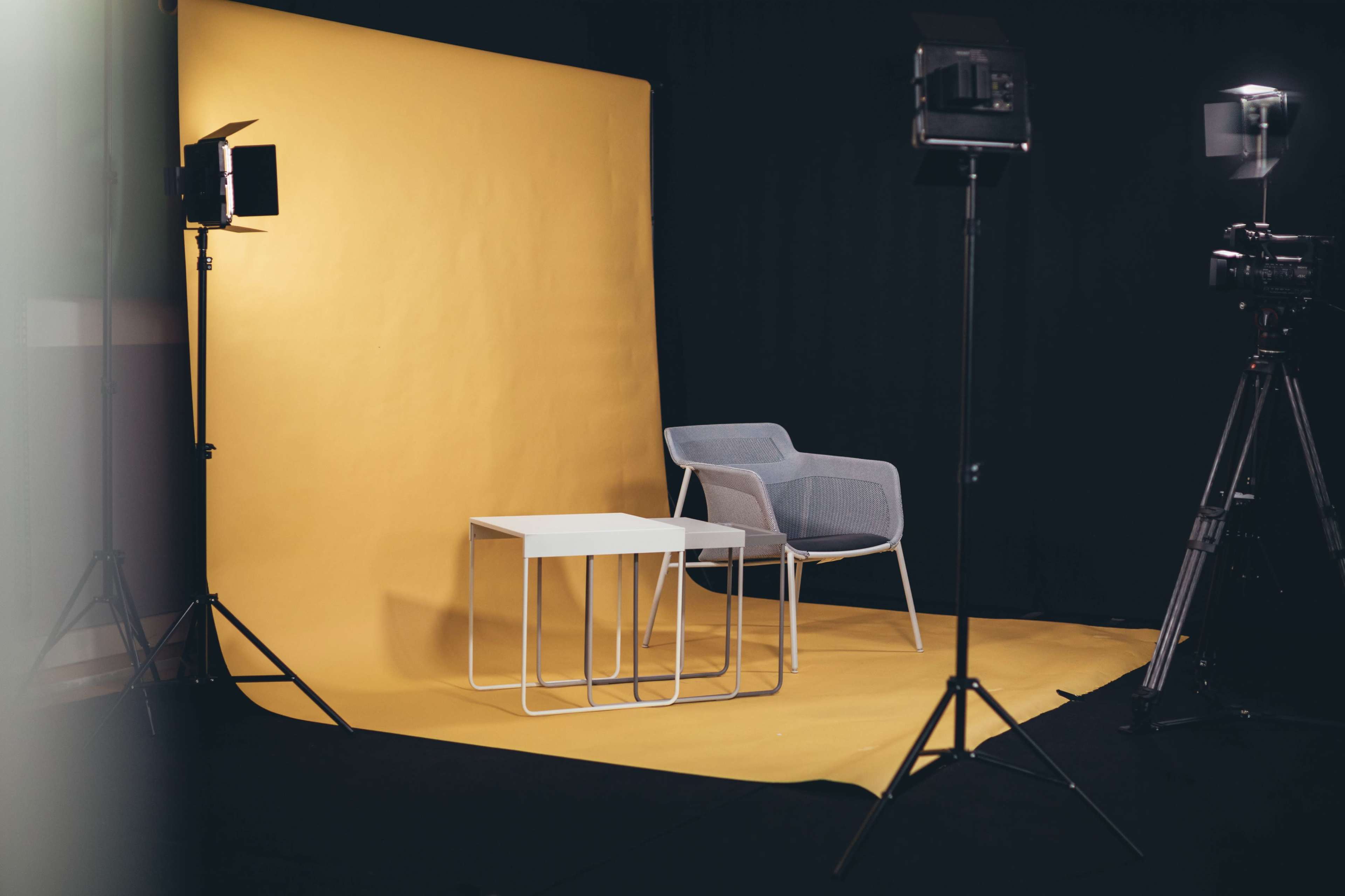 A gray chair and a white table are positioned in front of a yellow backdrop, illuminated by studio lights.