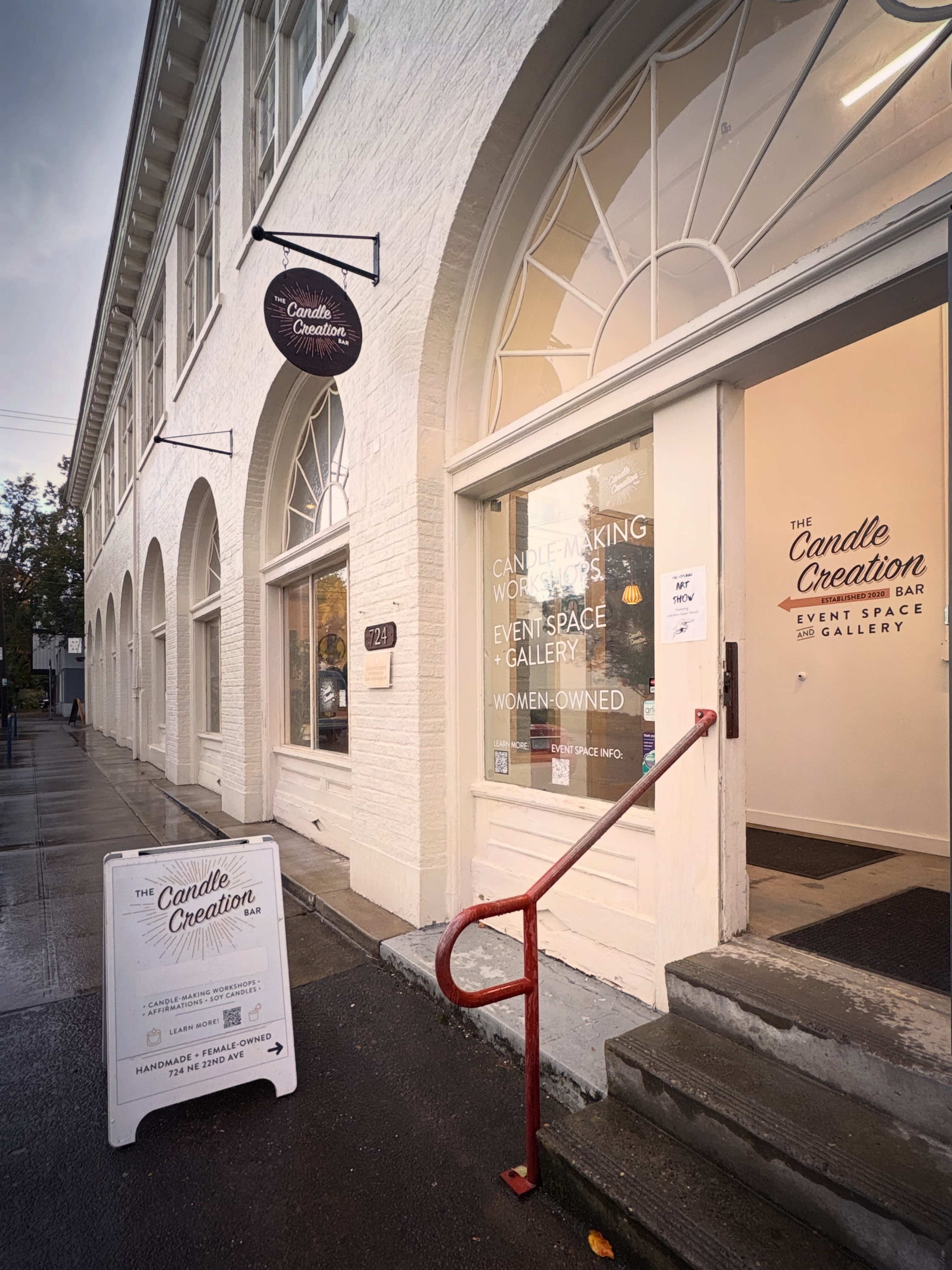 The image shows the exterior of "The Candle Creation," a candle-making workshop and event space, featuring a white building with large arched windows and a red handrail by the entrance.