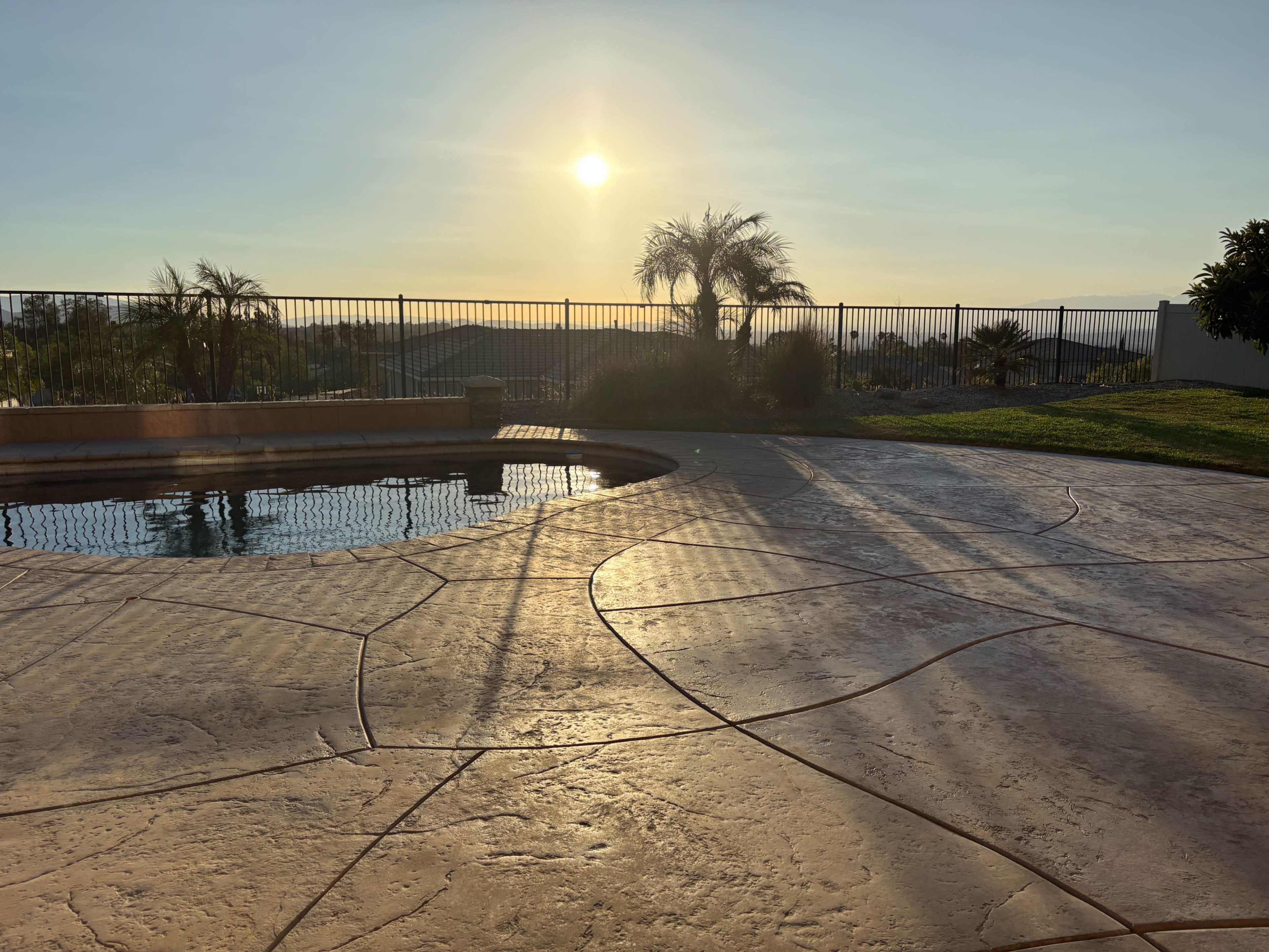 The image shows a serene pool area with a textured concrete surface, featuring a sunset against a backdrop of hills and palm trees.