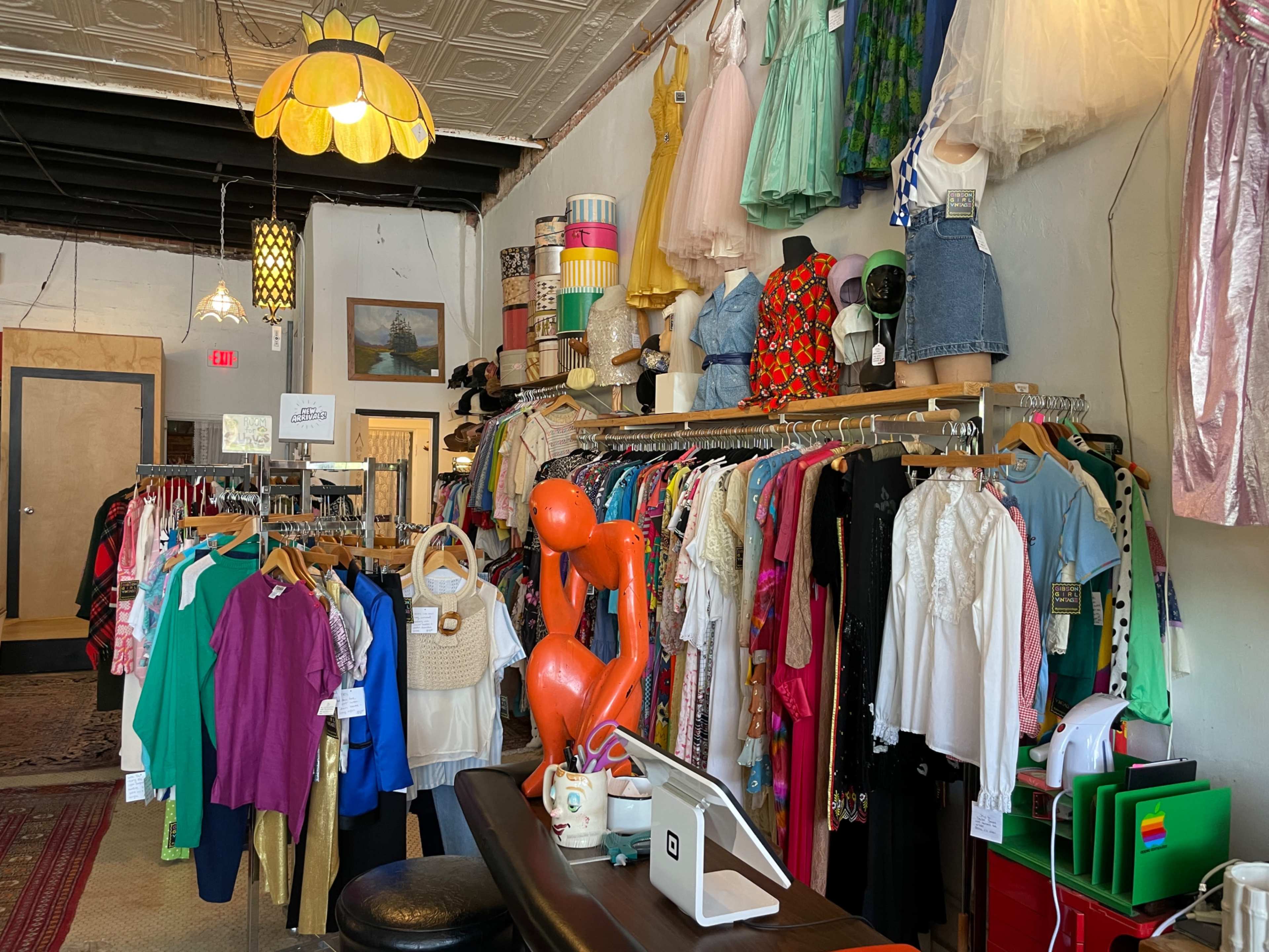 The interior of a colorful thrift shop displays racks of clothing and accessories along with various decorations overhead.