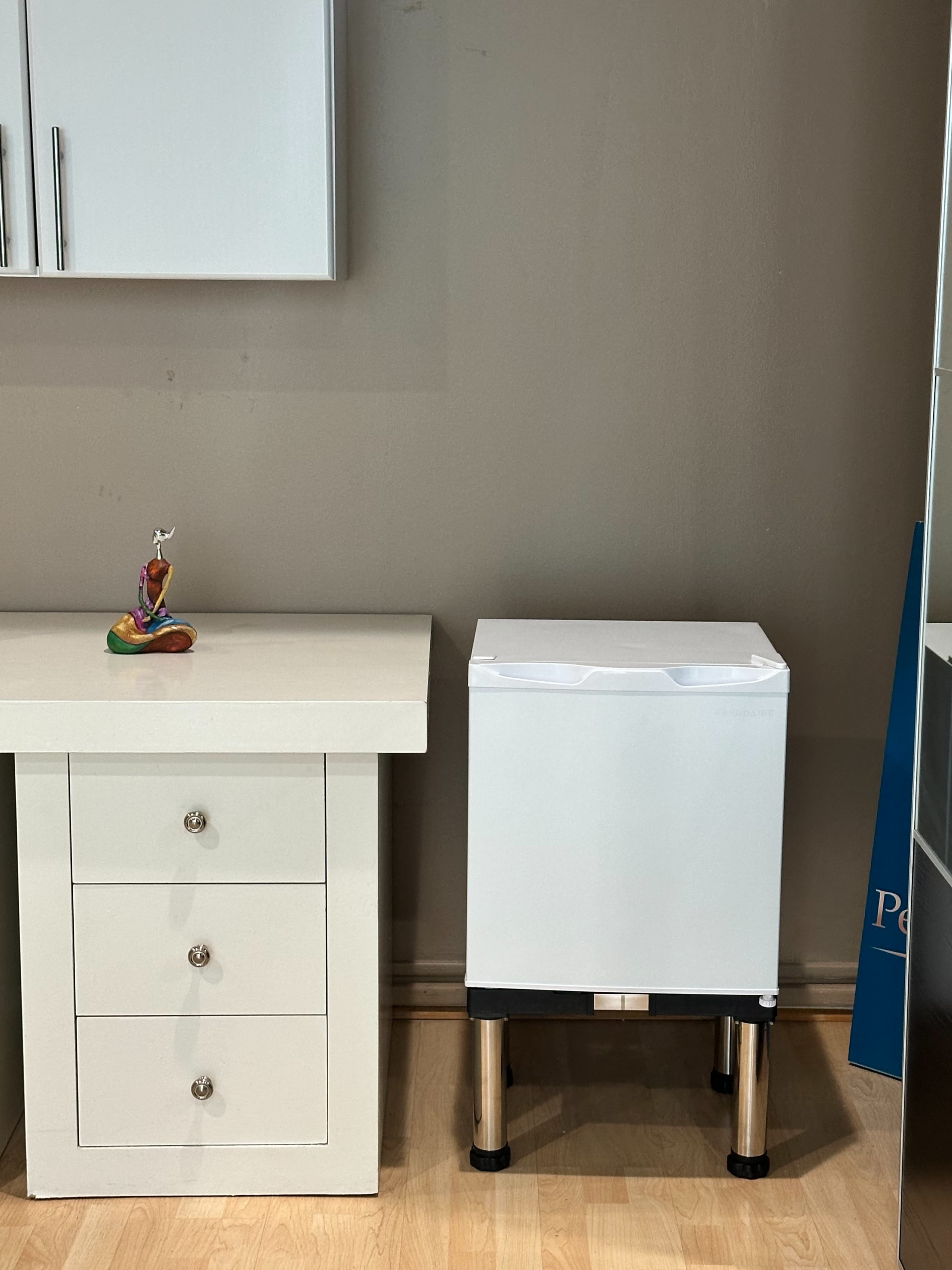 A white box-like appliance with metallic legs stands next to a desk with three drawers and a small decorative figure on top.