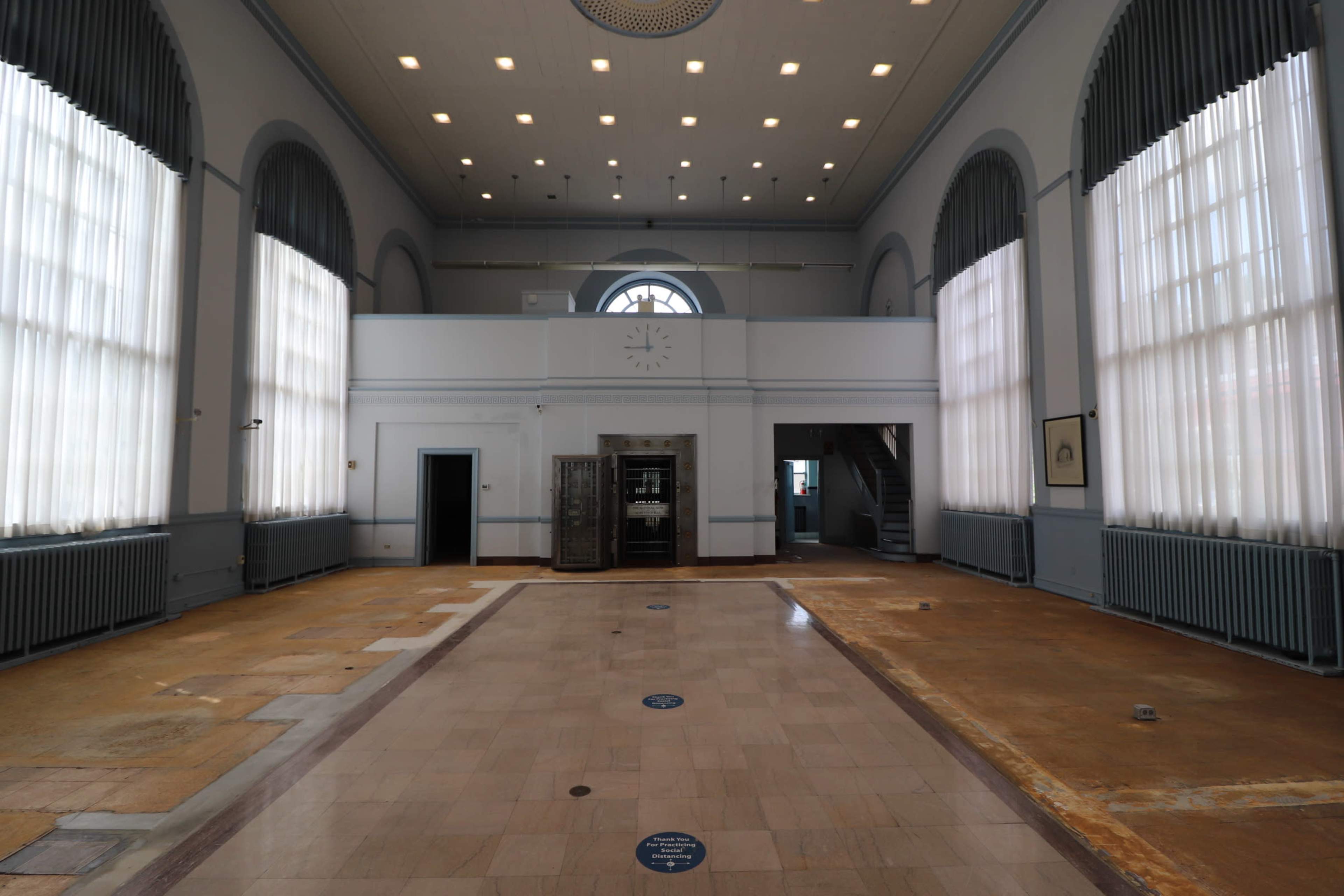 The image shows a spacious, empty room with high ceilings, large windows, and a central staircase at the far end.