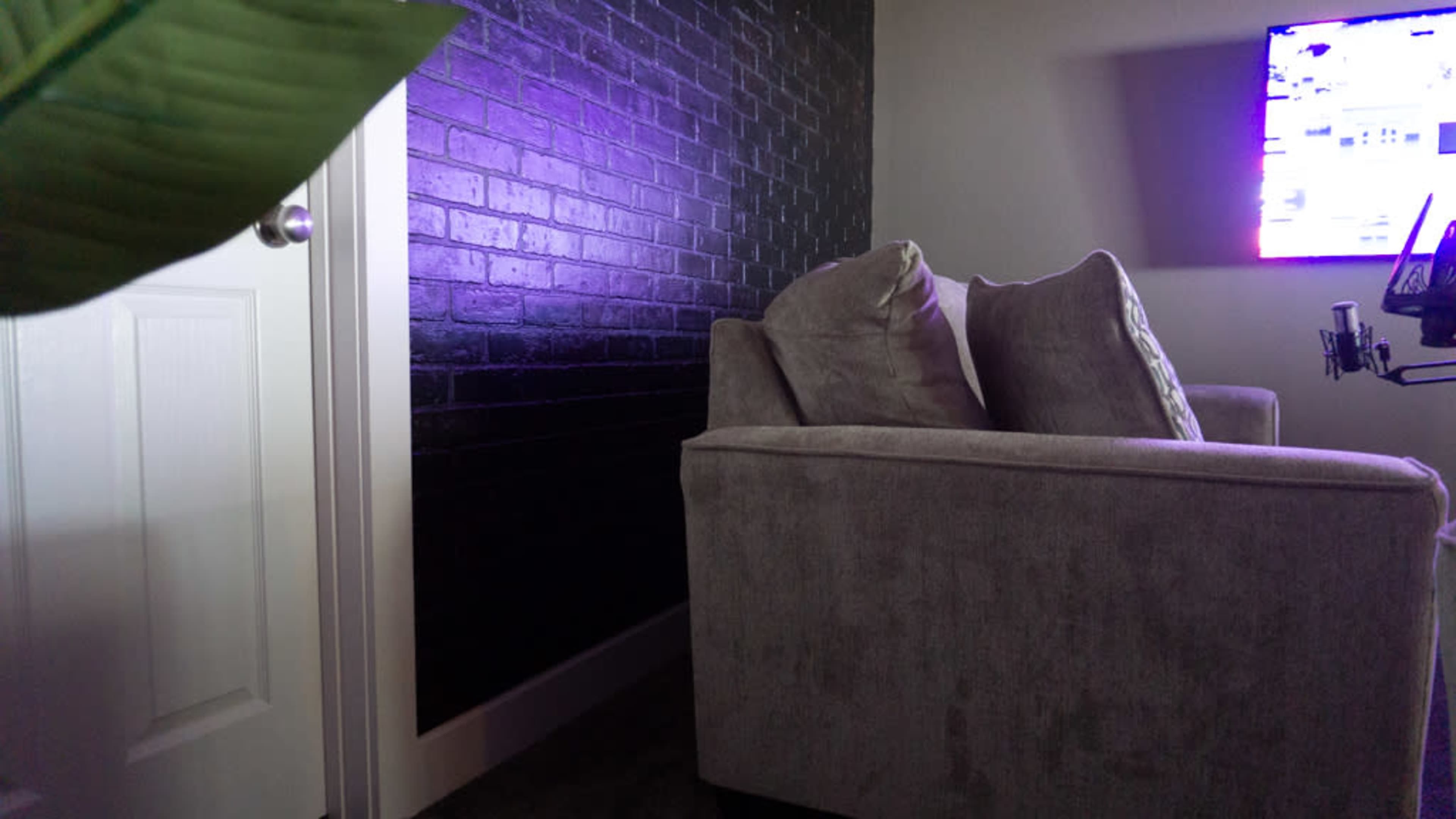 A gray couch is positioned near a door and a wall with a dark, textured surface, while a TV screen displays bright colors in the background.