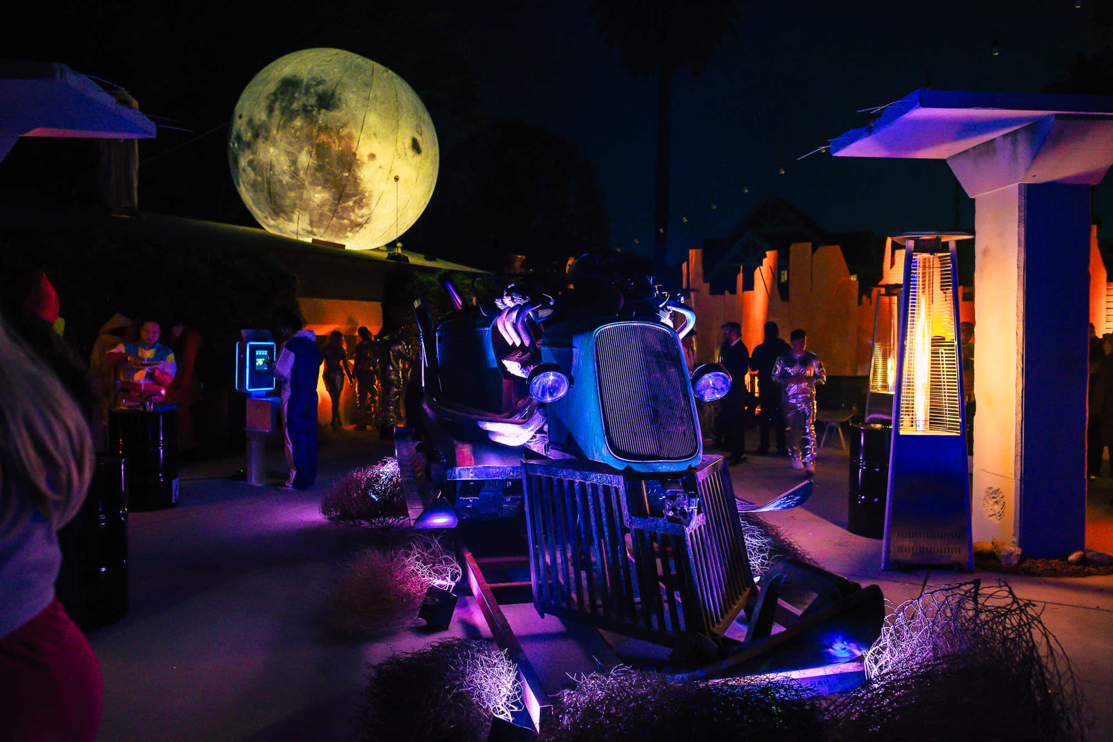 A vintage car is displayed in an outdoor setting illuminated by colorful lights, with a large moon backdrop and people mingling around.