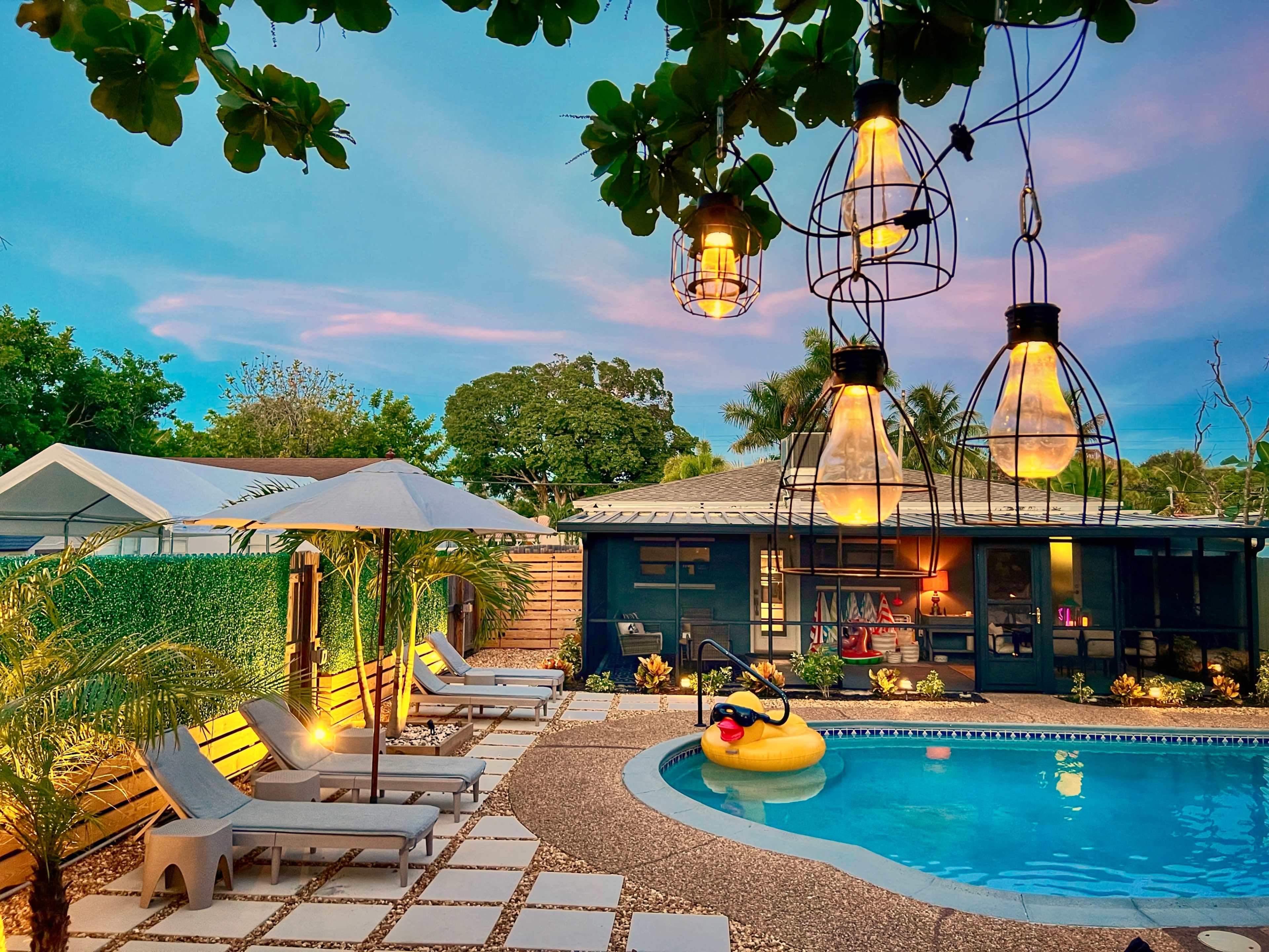 A well-lit backyard features a swimming pool surrounded by lounge chairs, a floating inflatable duck, and pendant lights hanging from overhead branches.